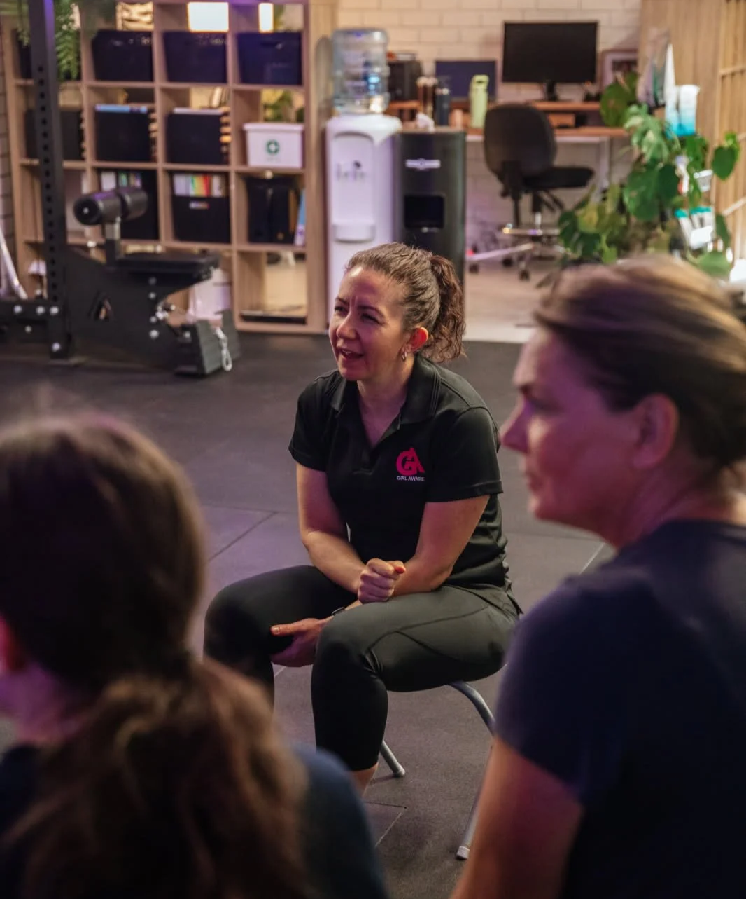 A woman with a ponytail, wearing a black 'Girl Awaken' shirt, is sitting on a chair and talking to a group in a modern office or fitness studio. In the background, there is a water cooler, a desk with a computer, bookshelves with storage boxes, and some plants.