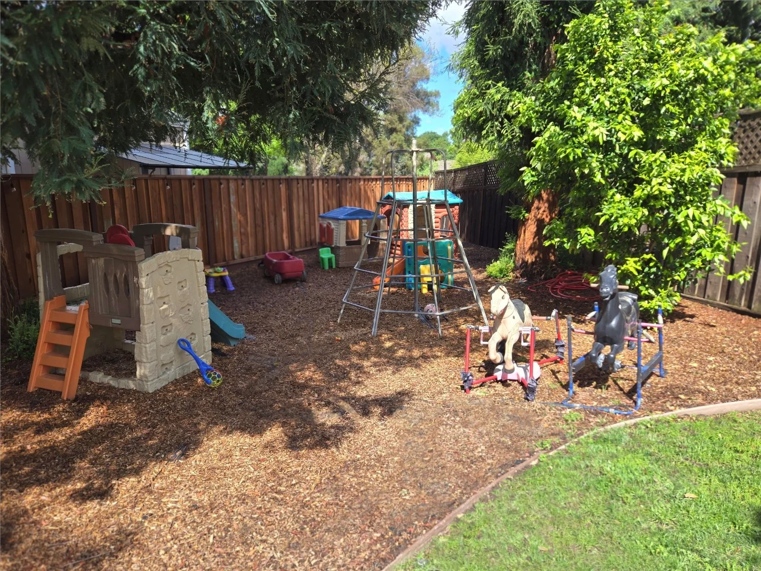 Children's backyard playground with a climbing structure, swings, and a playhouse, surrounded by trees and a wooden fence.