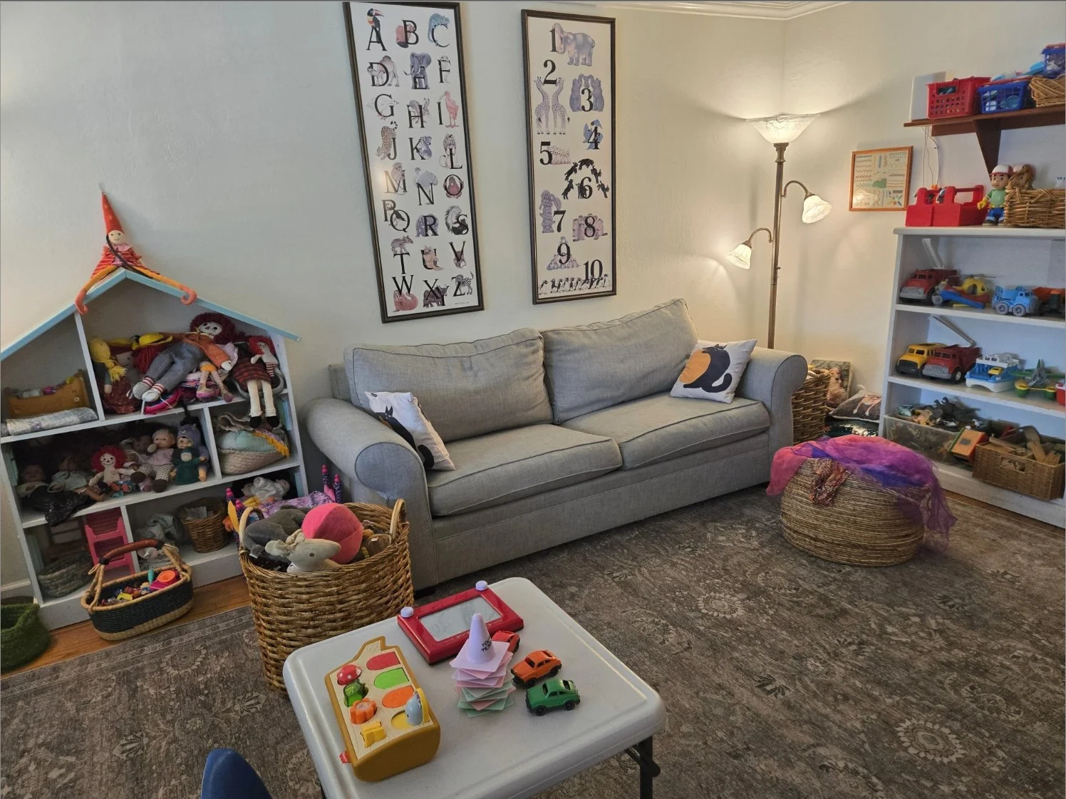 A cozy living room filled with toys and children's decor. There is a gray sofa with animal-themed pillows, bookshelf filled with toy cars and dolls, a small table with toy cars and a paper craft, framed alphabet and number charts on the wall, a standing lamp with two lights, and baskets filled with toys on the floor.