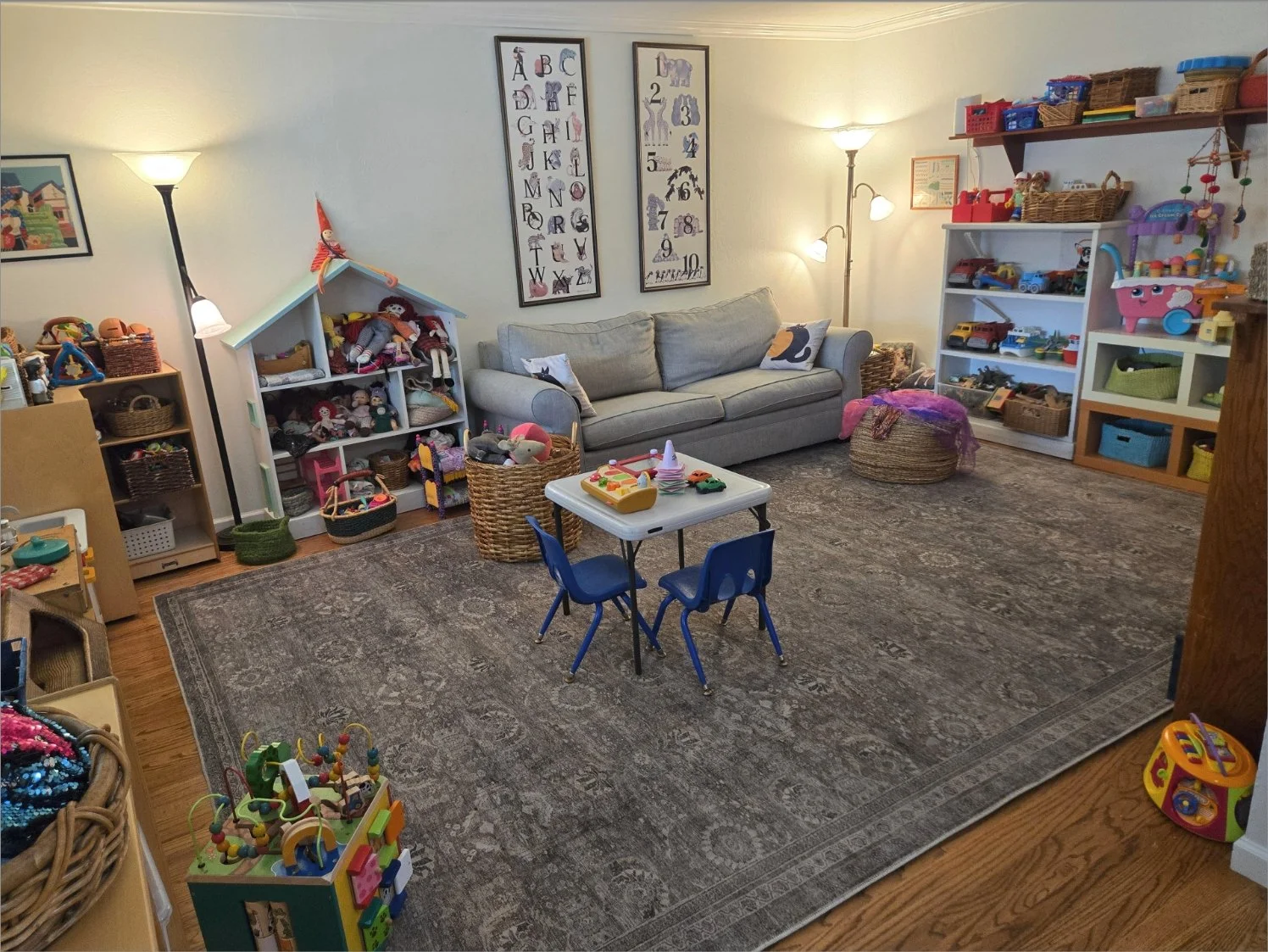 A cozy children's playroom with a gray sofa, children's toys, and stuffed animals on shelves. There are colorful chairs and a small table in the center, surrounded by toy storage and a large area rug. The room is warmly lit with lamps and wall decor.