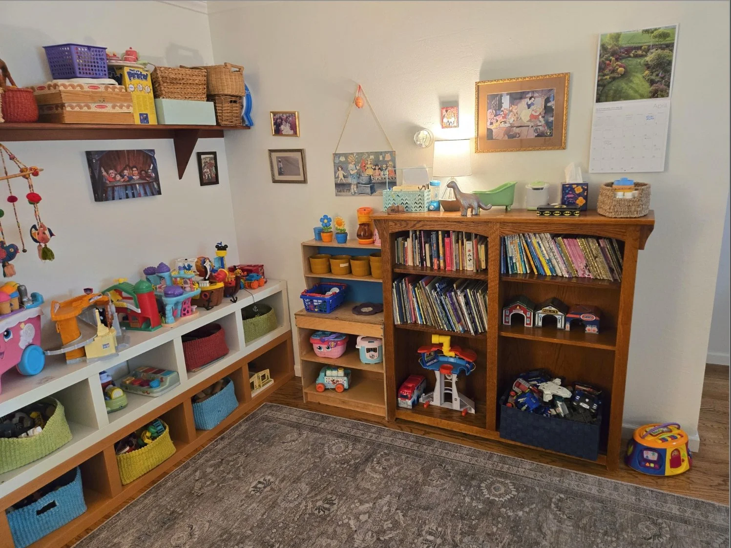 A corner of a child's playroom with shelves filled with toys, books, and children's decorations. There is a bookshelf, a small bookcase, a wall shelf with framed pictures, and various toys on top and inside the storage units. A wall calendar and wall art are also visible.
