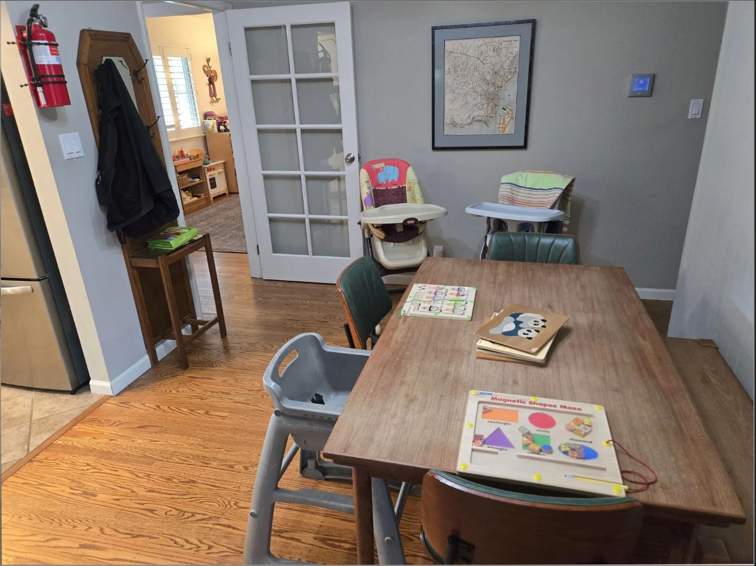 A dining area with a wooden table surrounded by green and wood chairs, a children's puzzle and folder on the table, and a high chair nearby. There's a gray wall with a framed map, a wooden coat rack with a bag hanging, and a doorway leading to another room with toys. A child car seat is against the wall, and a small wooden stand with books and a green object is next to the coat rack. The room has hardwood floors.