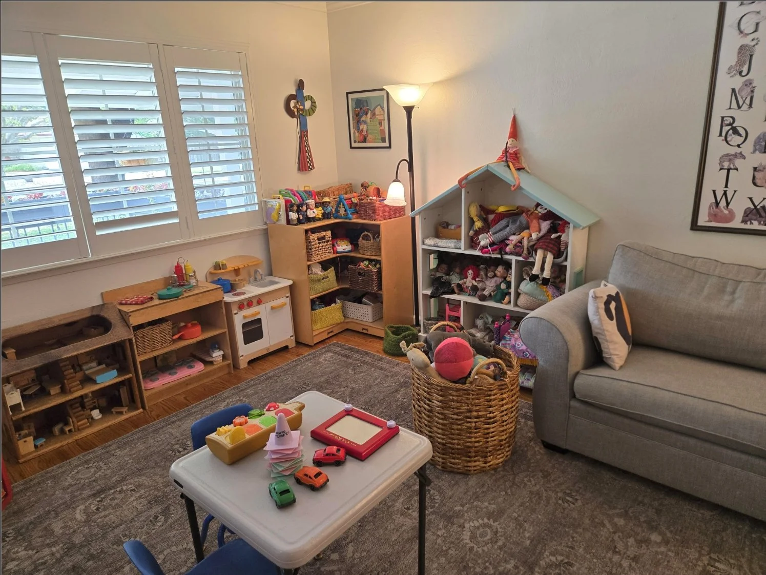 Children's playroom with toy kitchen, shelves filled with dolls and toys, a small table with toy cars, and a sofa, all near windows with white shutters.