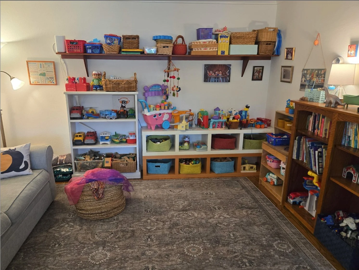 A cozy children's playroom with white and wooden shelves filled with toys, books, and baskets. A gray couch with a decorative pillow is on the left, and a woven basket with a pink scarf sits on the floor in front. The walls feature framed pictures, and a lamp is on the far right.