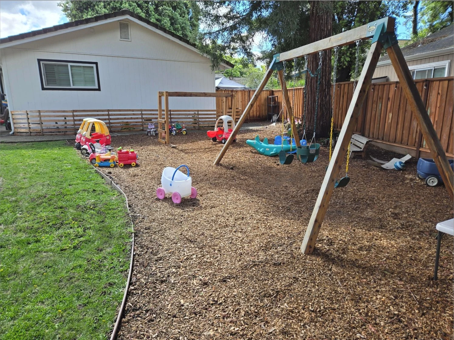 Rear backyard with a children's play area featuring a tire swing set, ride-on toys, small plastic houses, a sandbox, and a wooden fence, with a house in the background and trees lining the yard.