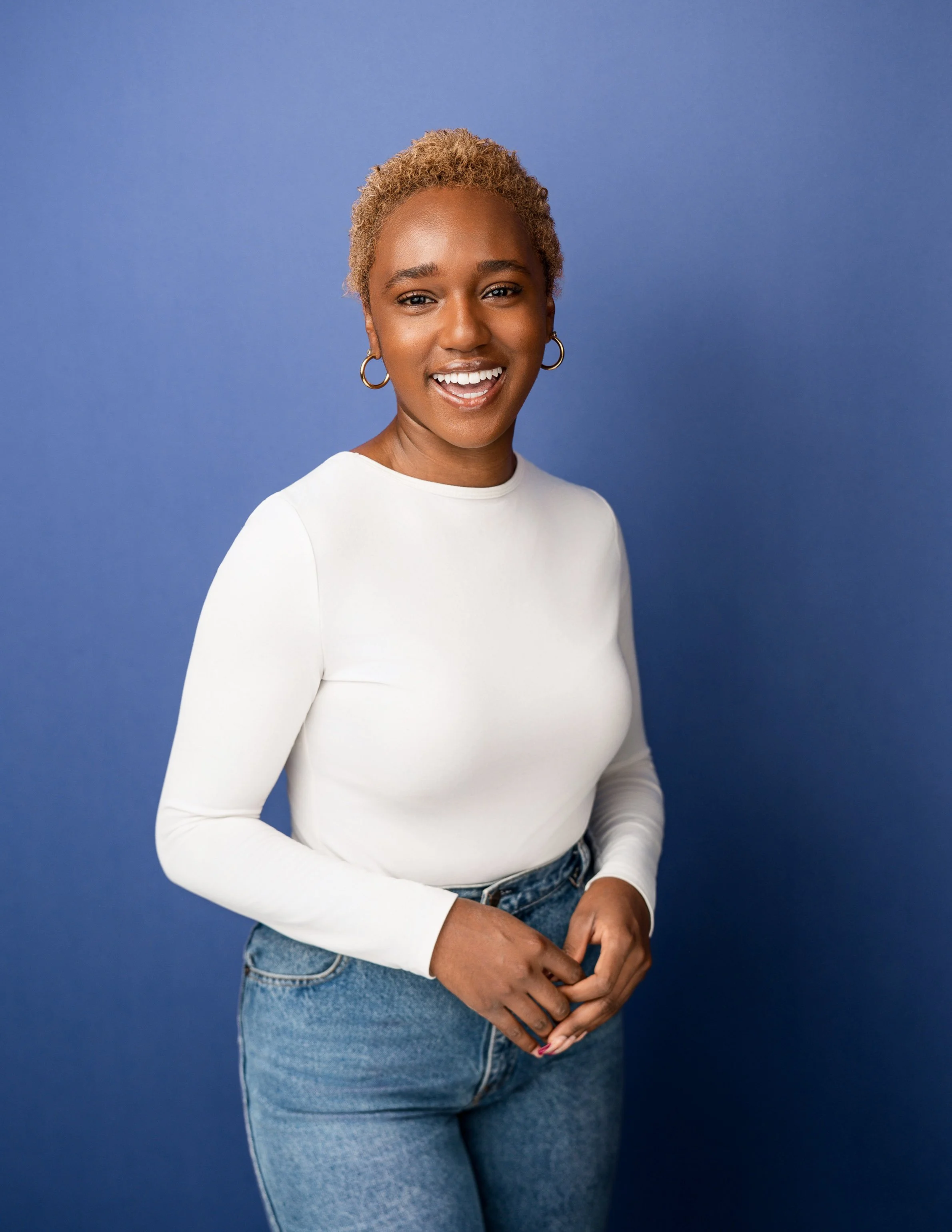 A woman with short curly blonde hair smiling, wearing a white long-sleeve top and blue jeans, standing against a blue background.