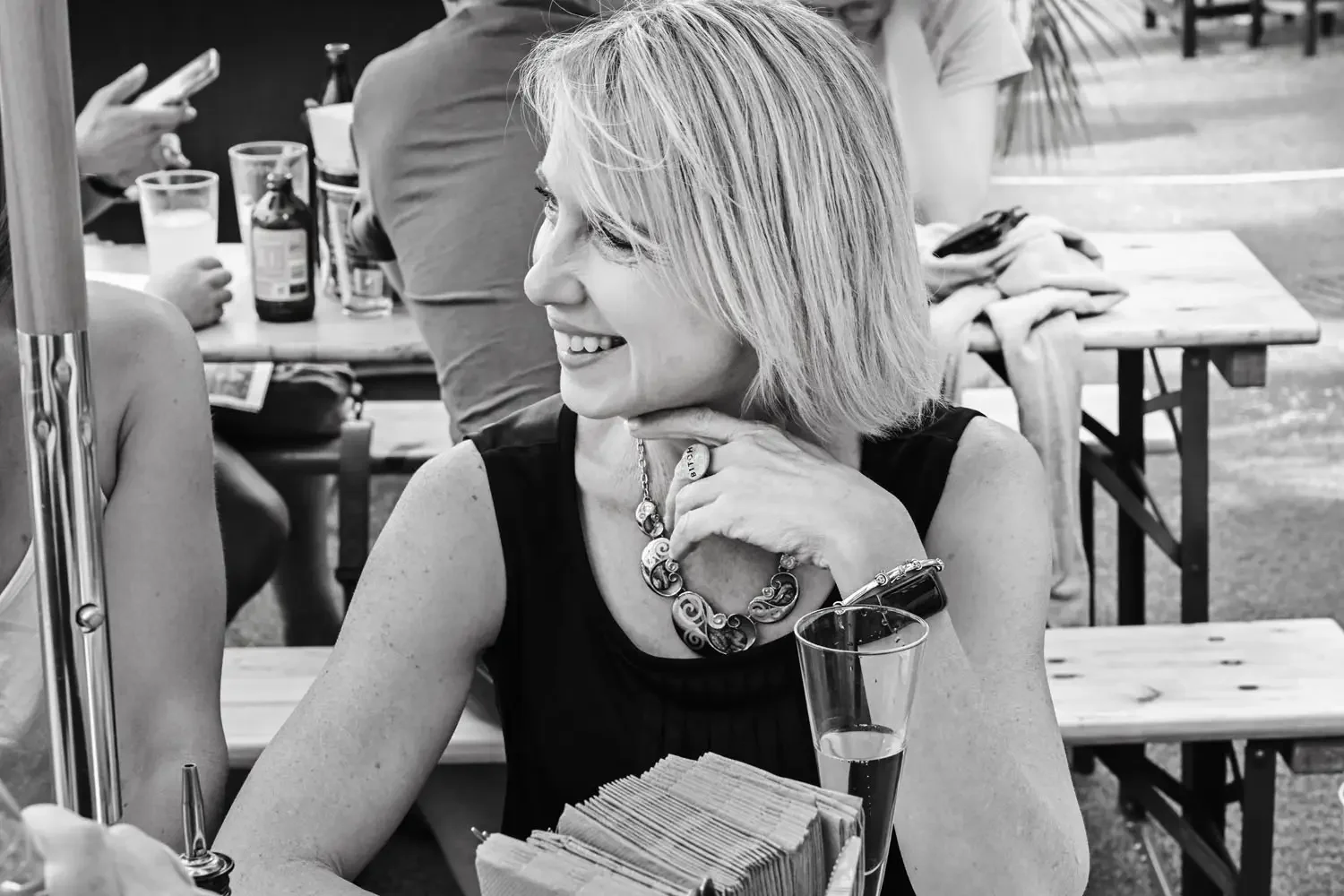A woman with a short blonde haircut, smiling and sitting at an outdoor table, wearing a sleeveless black top, a large statement necklace, and a bracelet. There are drinks and other people at tables in the background.