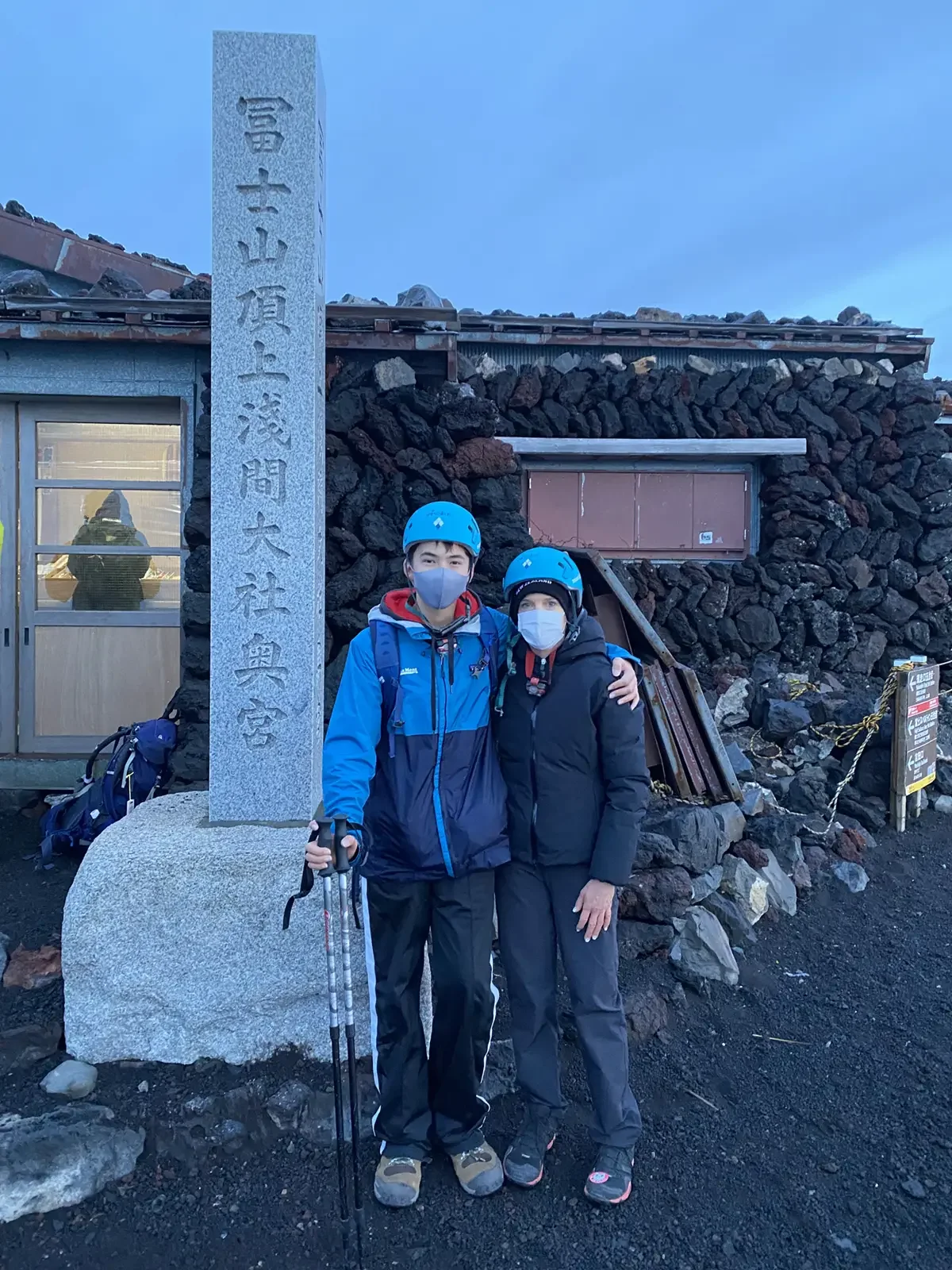 Two people in outdoor winter gear and helmets standing at Mount Fuji's 3,776-meter marker at the summit, smiling with arms around each other, wearing face masks. Behind them are stacked rocks, a small building, and a stone monument with Japanese inscriptions, all under a clear blue sky.