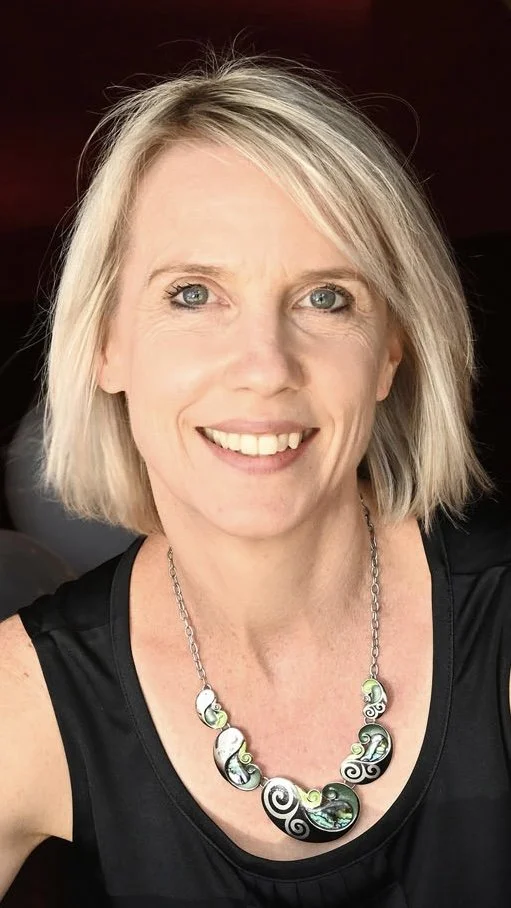 A smiling woman with blonde hair and blue eyes wearing a black top and a colorful necklace.