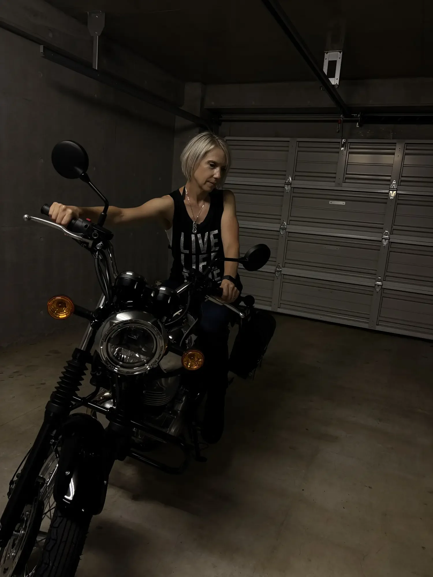 A woman with short blonde hair sitting on a black motorcycle in a dimly lit garage, wearing a black tank top with silver lettering.