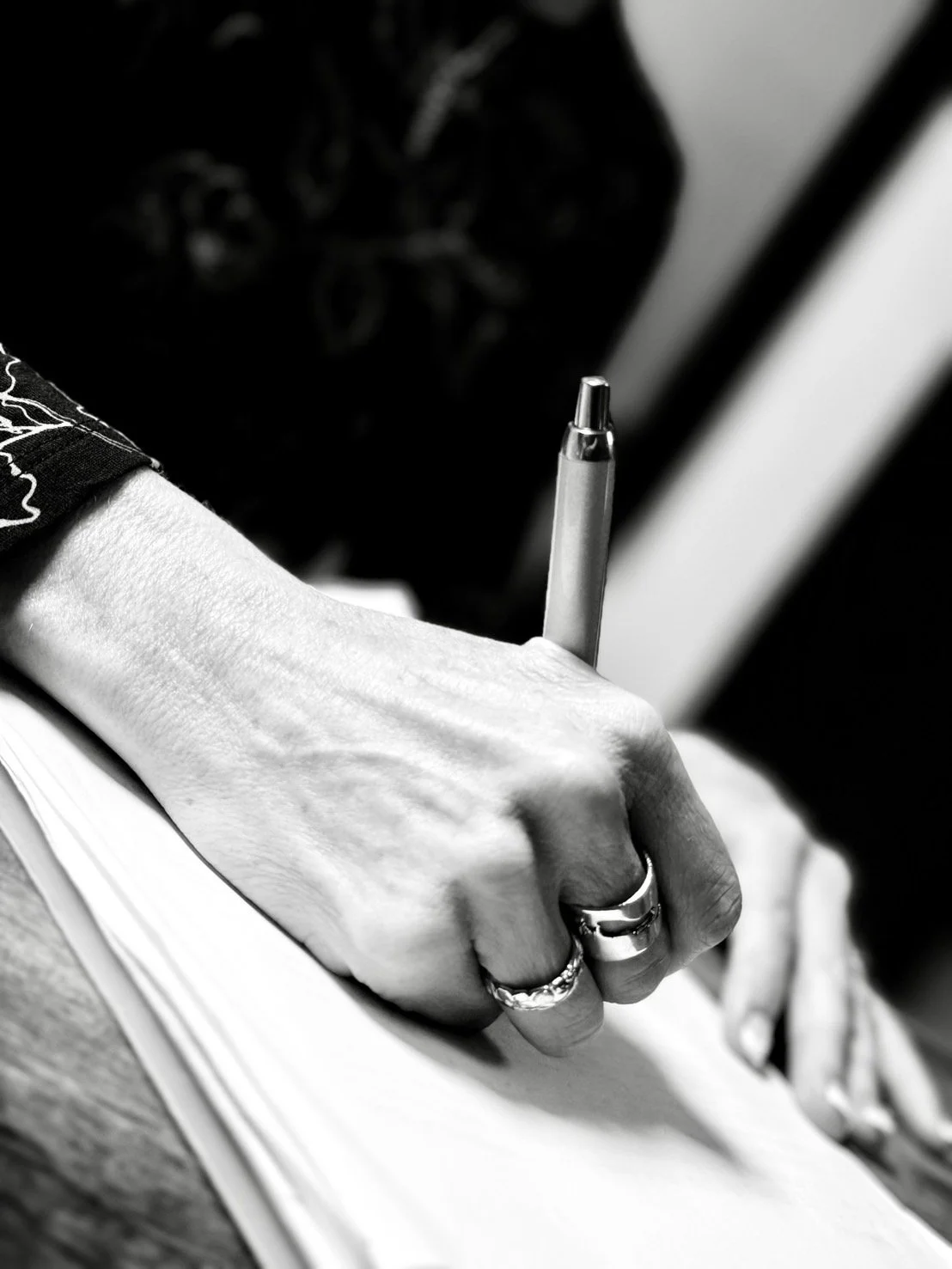 A person's hand is holding and writing with a pen on paper, showing rings on fingers and a tattoo on the wrist, in black and white.