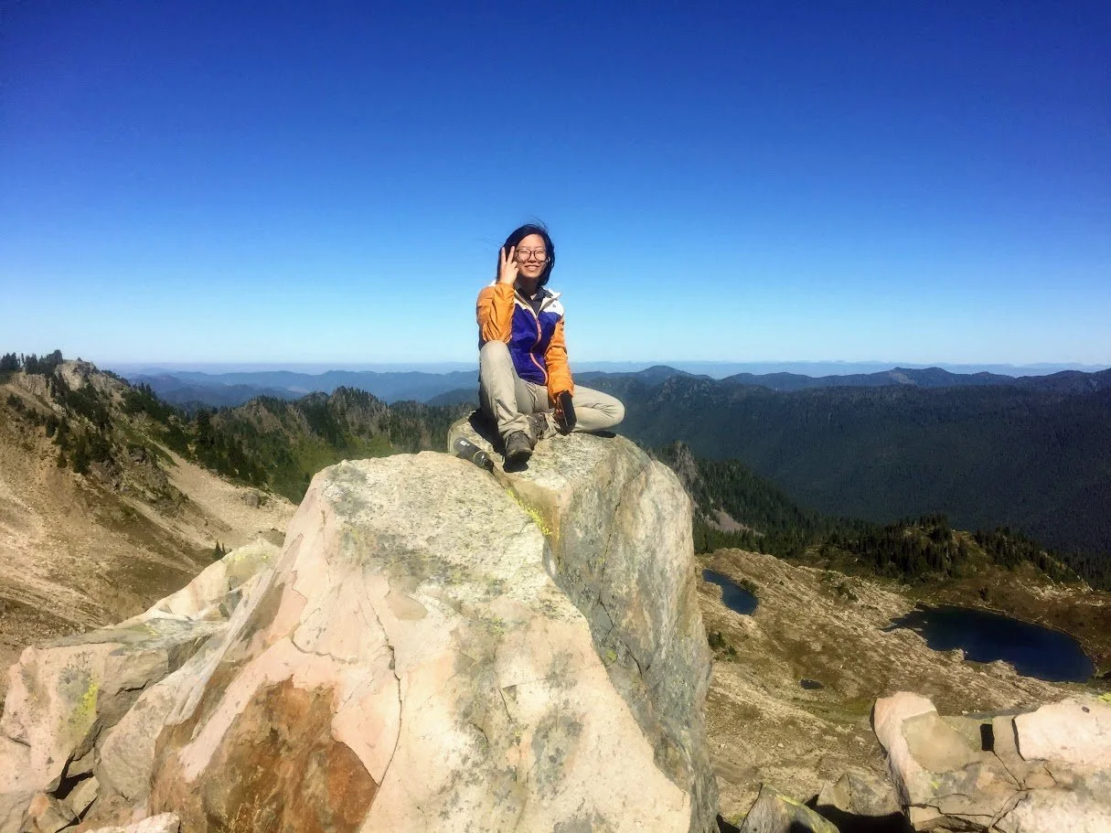 A woman sitting on a large rock at a mountain summit with a vast mountain range and lakes in the background, under a clear blue sky.