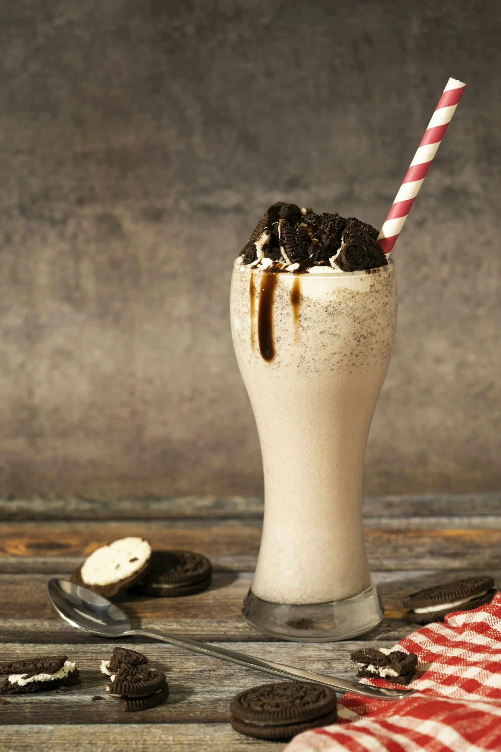 Batido de leche con helado, Oreo y caramelo en un vaso alto, con pajilla roja y blanca, en una superficie de madera con galletas Oreo y un cucharador.