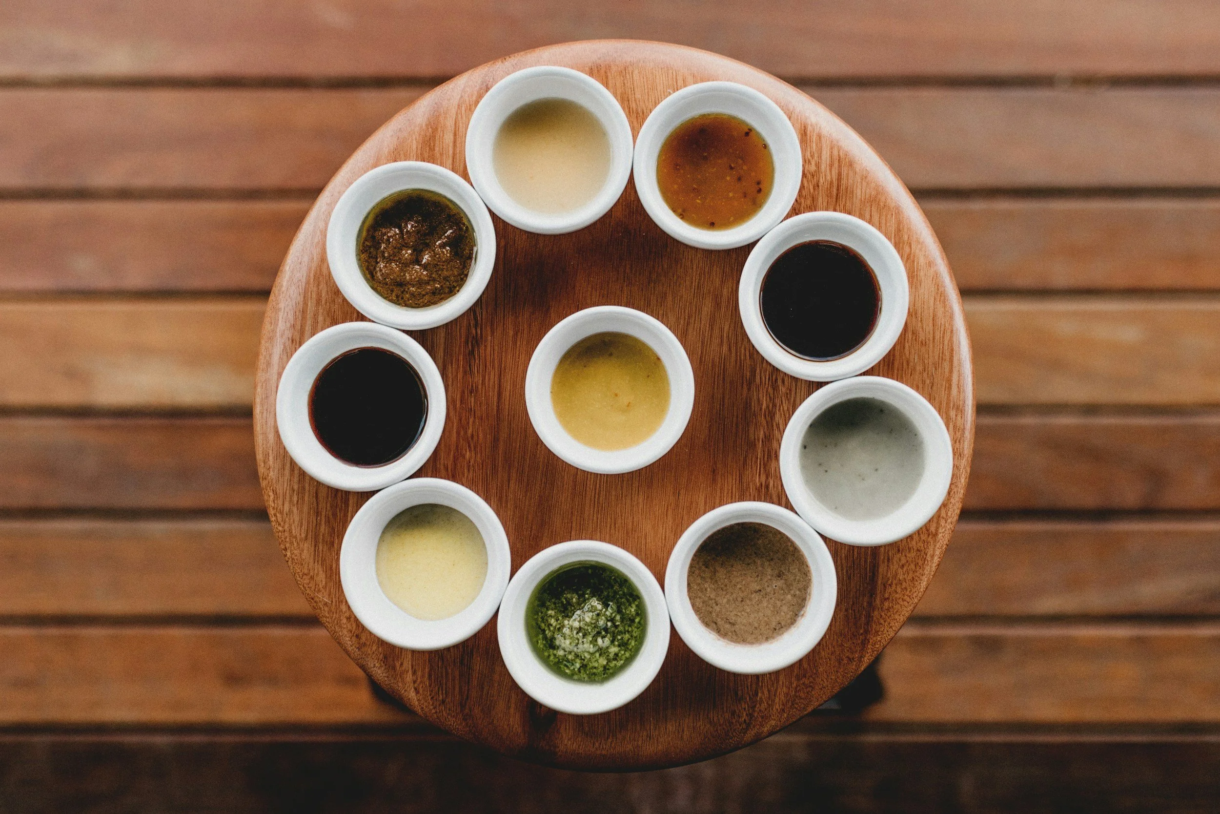 Muestra una variedad de salsas o aderezos en pequeños recipientes blancos sobre una mesa de madera.