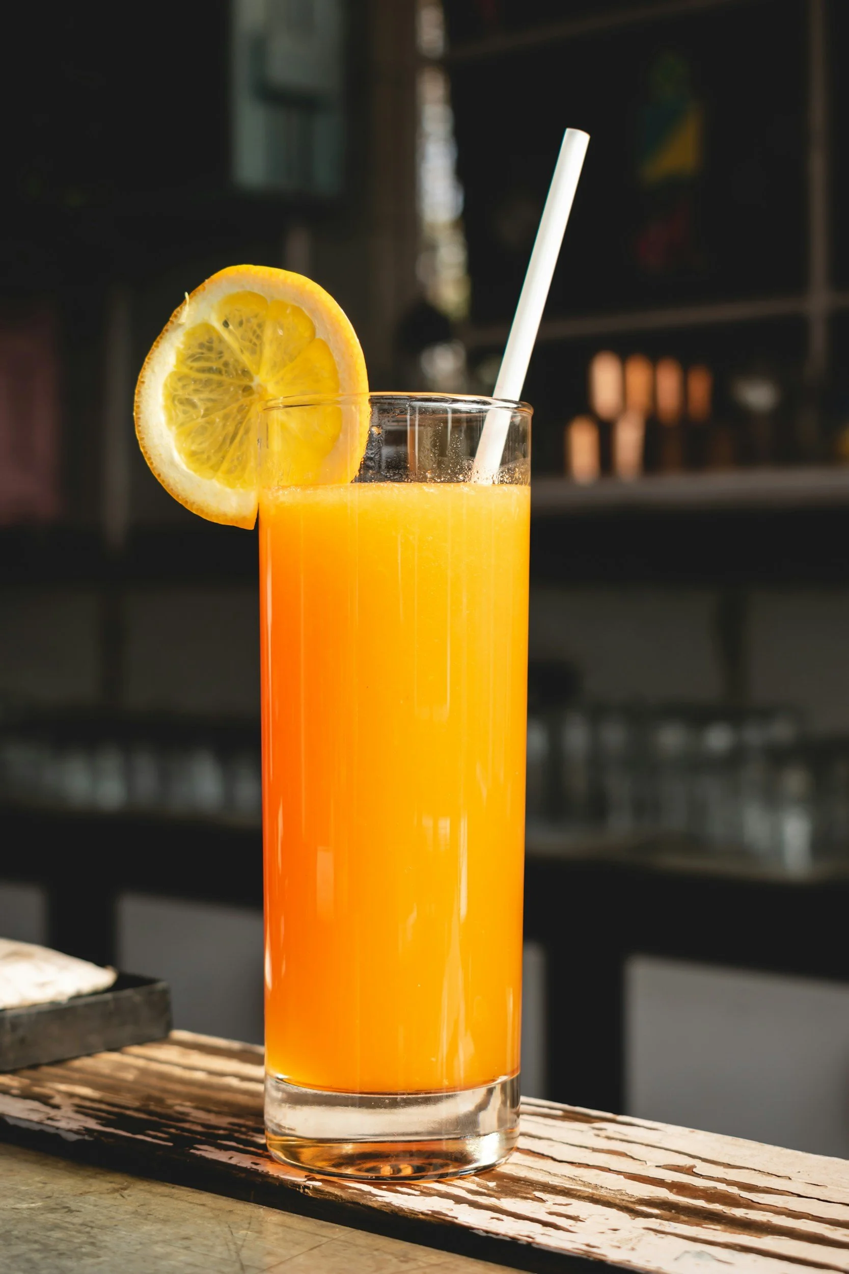 Vaso alto con jugo de naranja, decorado con una rodaja de limón en el borde y una pajilla blanca, sobre una mesa de madera.