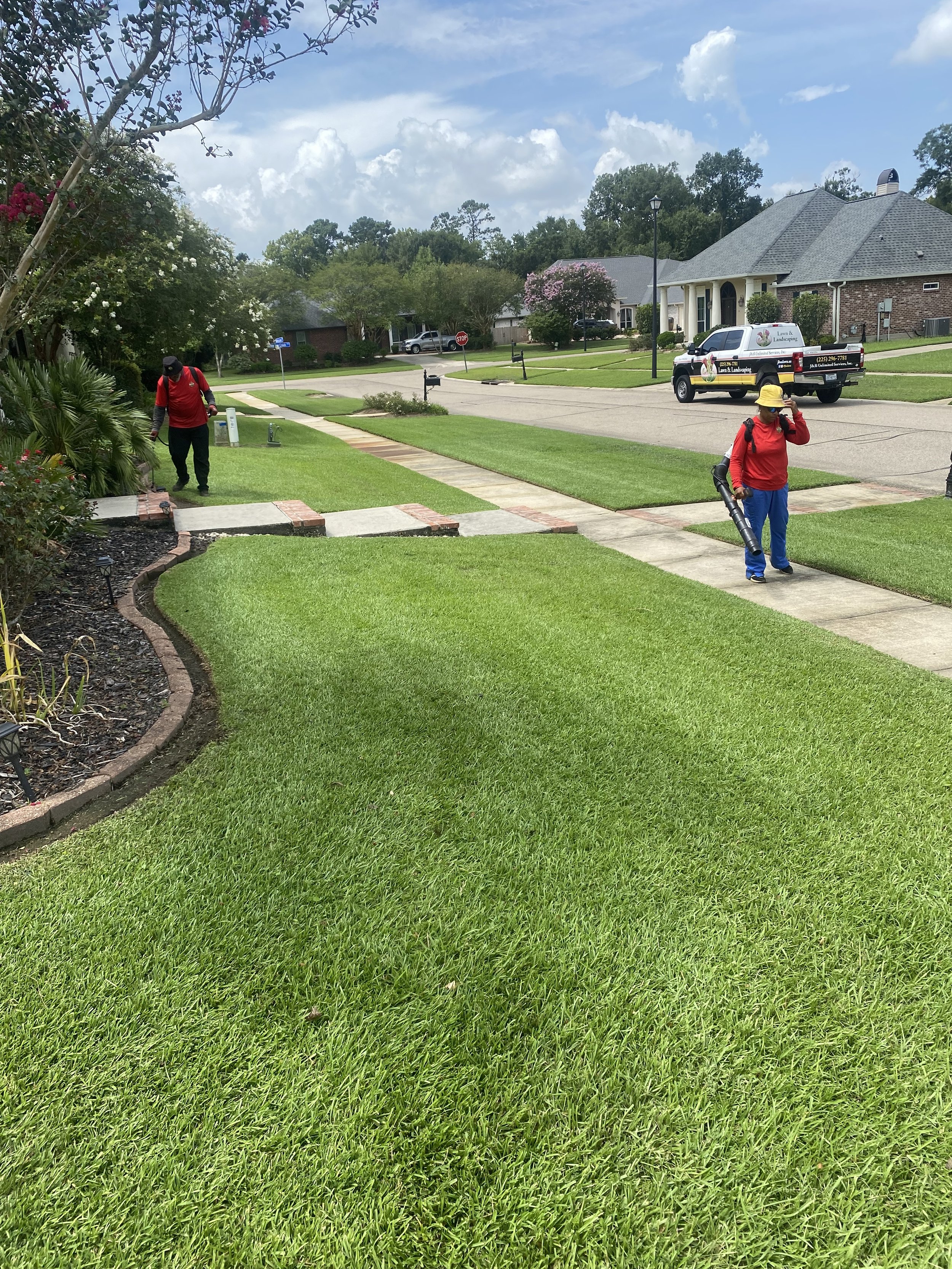Two landscaping workers, dressed in red shirts and blue pants, are working on a well-maintained front yard with lush green grass. One is using a leaf blower, while the other is walking along the sidewalk, adjusting his hat.