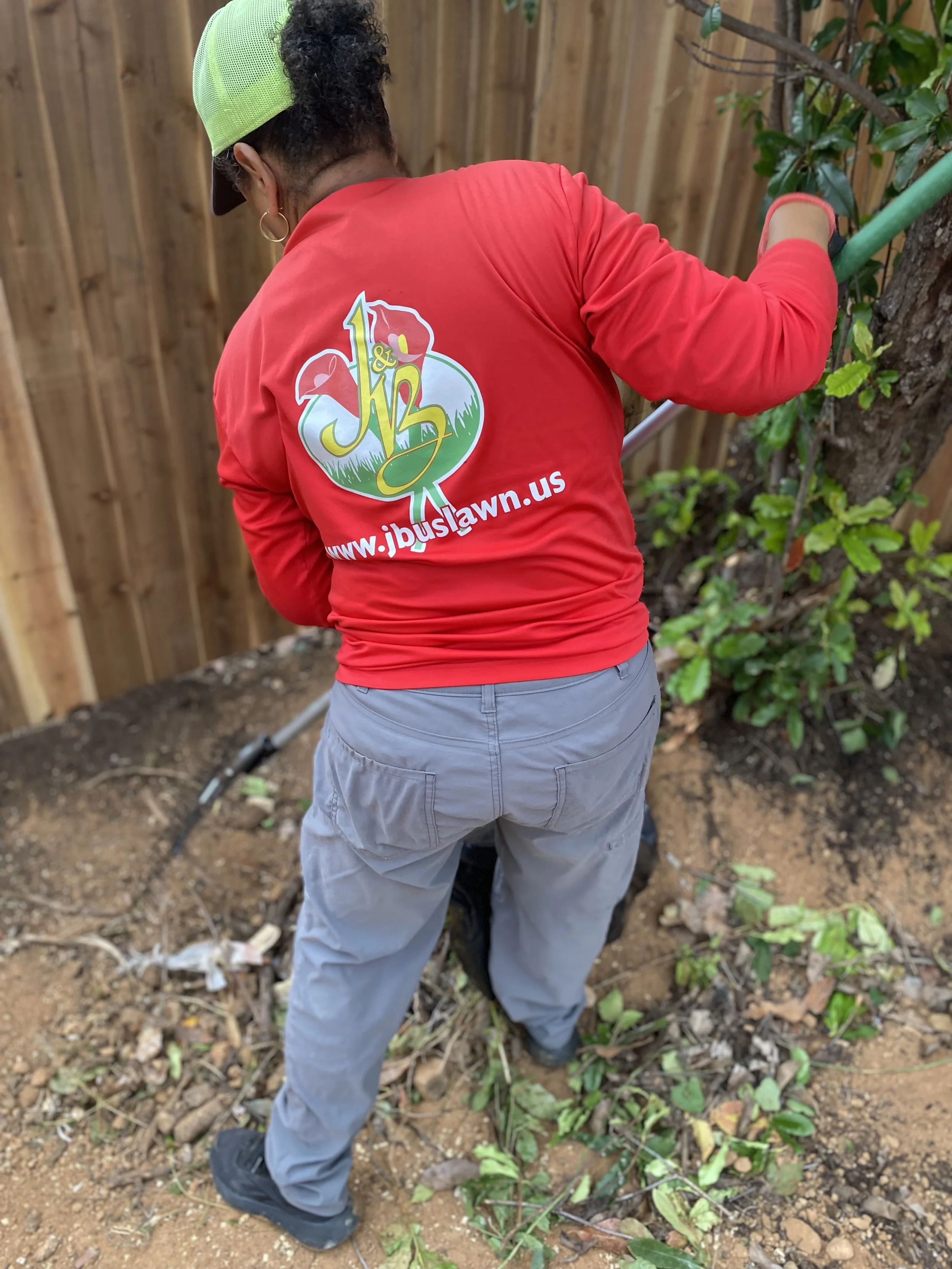 A J&B Landscaping crew member is raking up shrub debris in a garden or yard, wearing a J&B Lawn & Landscaping shirt with gray pants, a green cap, with a wooden fence and green foliage in the background.