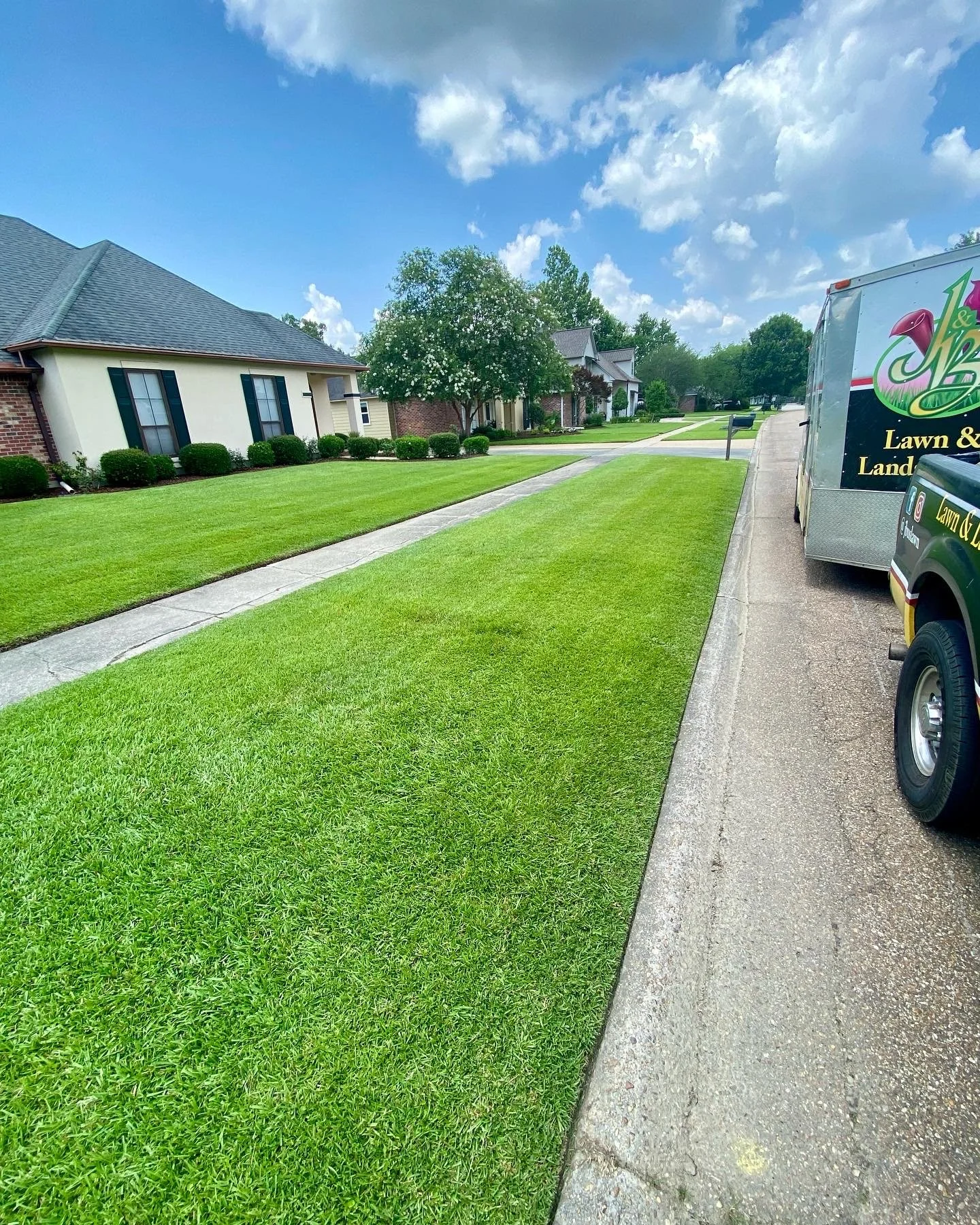 Nicely mowed residential lawn with healthy green a vibrant grass. Clear blue and partly cloudy sky with light tan house and a J&B Lawn & Landscaping truck on the street.