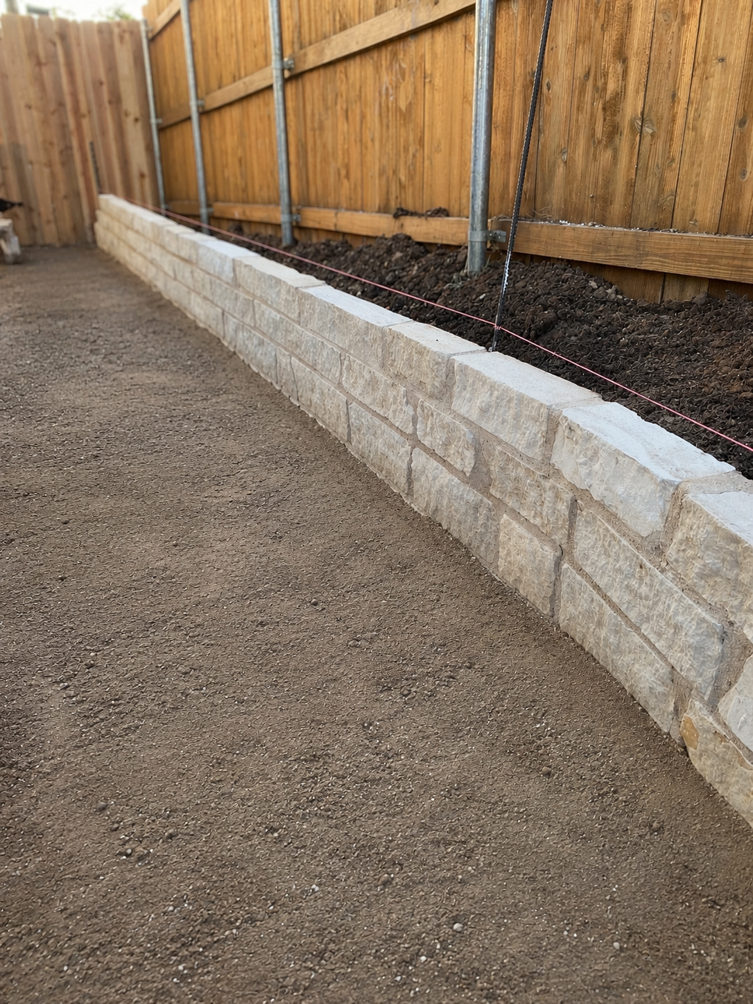 Cleaned retaining wall and graded soil area along wood fence during hardscaping project.