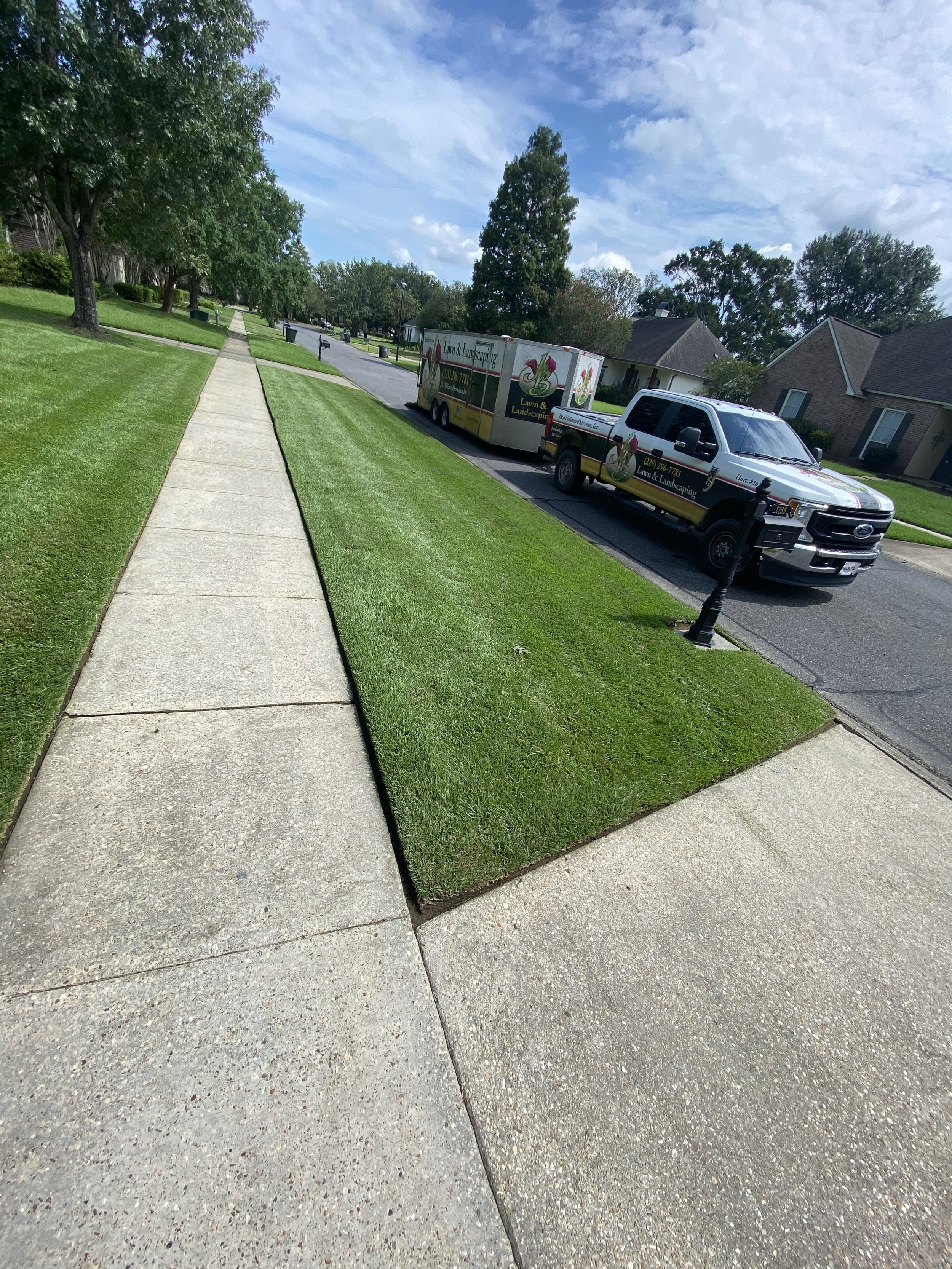 A suburban street with a clean edged sidewalk, green lawns, trees, and a J&B Lawn & Landscaping truck parked on the street. The sky is partly cloudy.