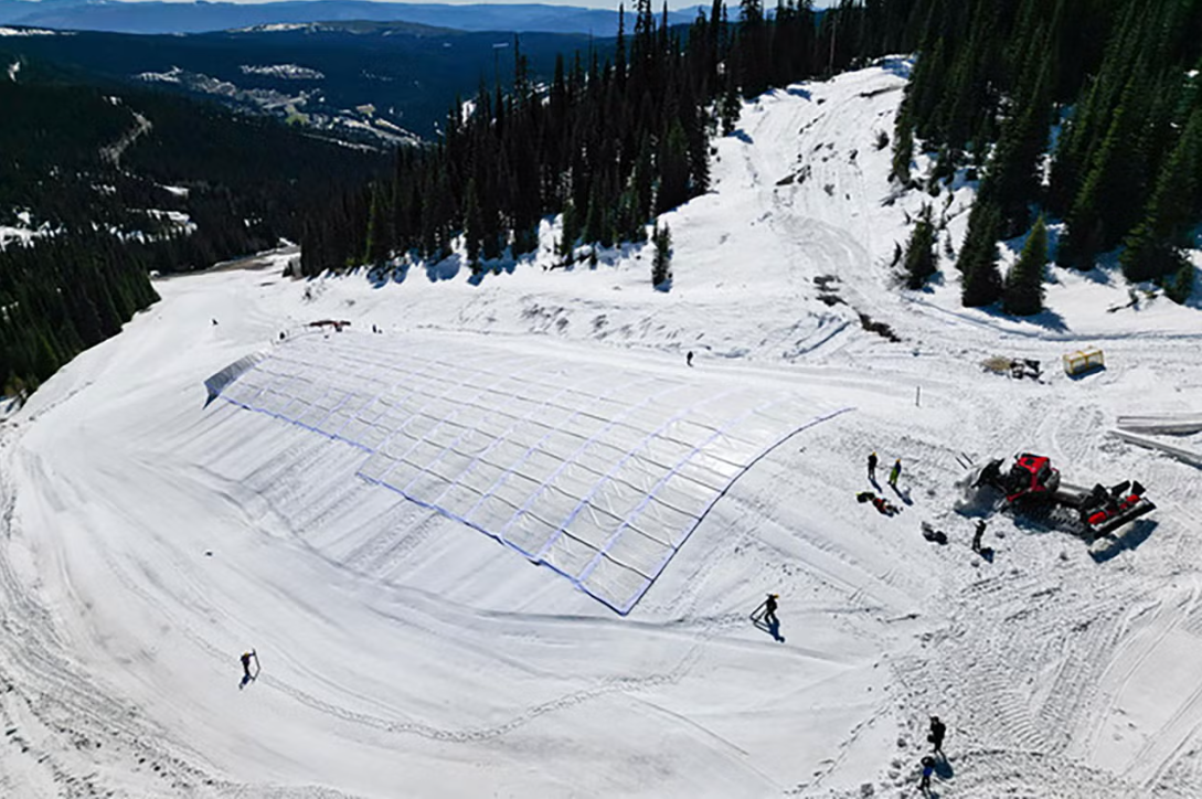 Snow-covered mountainside with workers installing a temporary shelter or greenhouse structure on the snow. Trees and distant mountain ranges are visible in the background.
