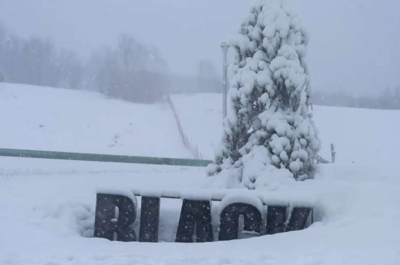 A snow-covered landscape with a snow-laden tree, a fence post, and a fallen sign with the partial word 'RACING' visible.