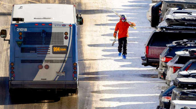 A person in a red jacket and black pants walking on a snowy street with parked cars on the right side and a bus in front, snow melting on the ground.