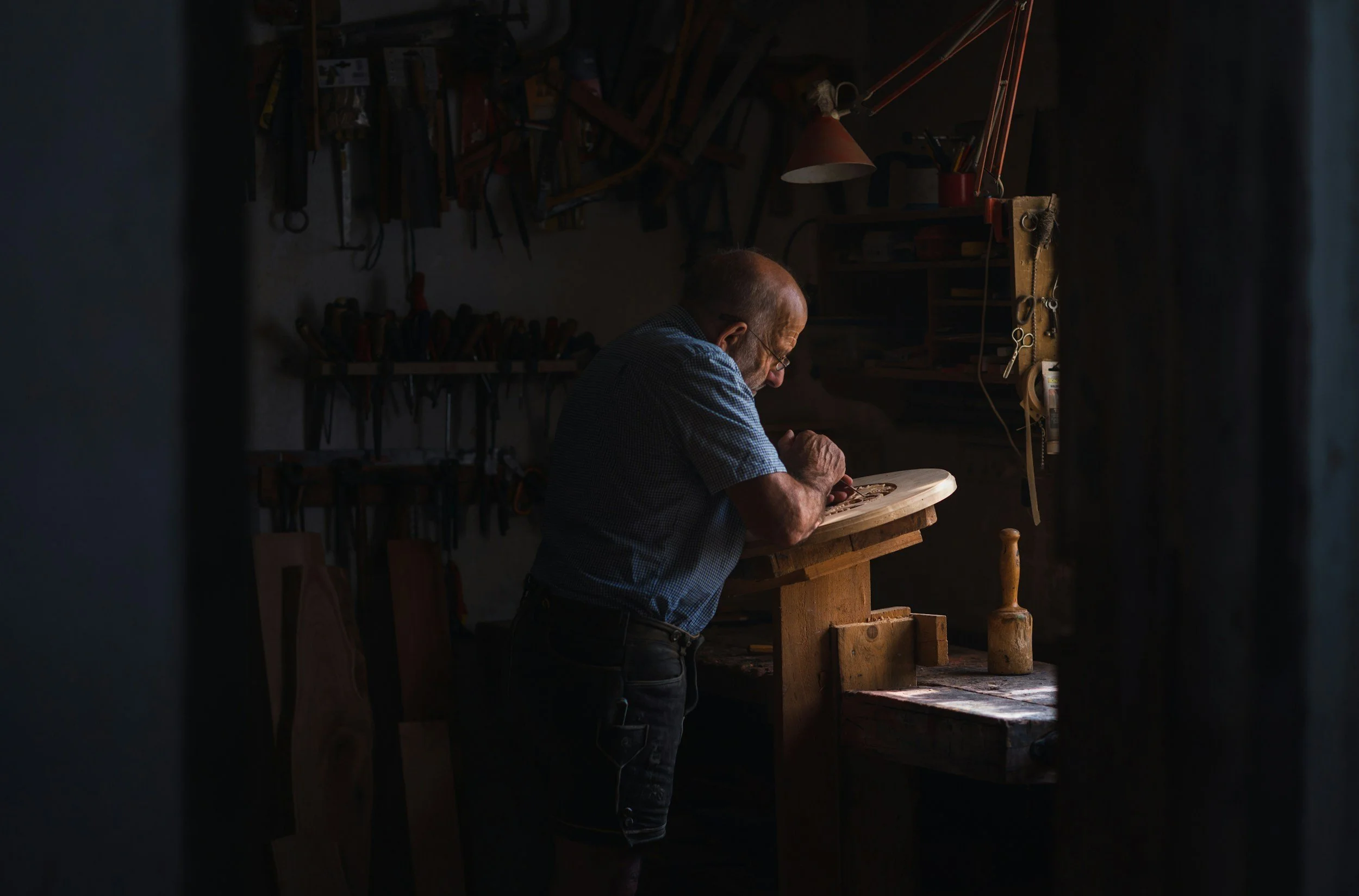 A man working in a woodworking workshop, focused on carving or shaping a piece of wood at a workbench, surrounded by tools hanging on the wall.