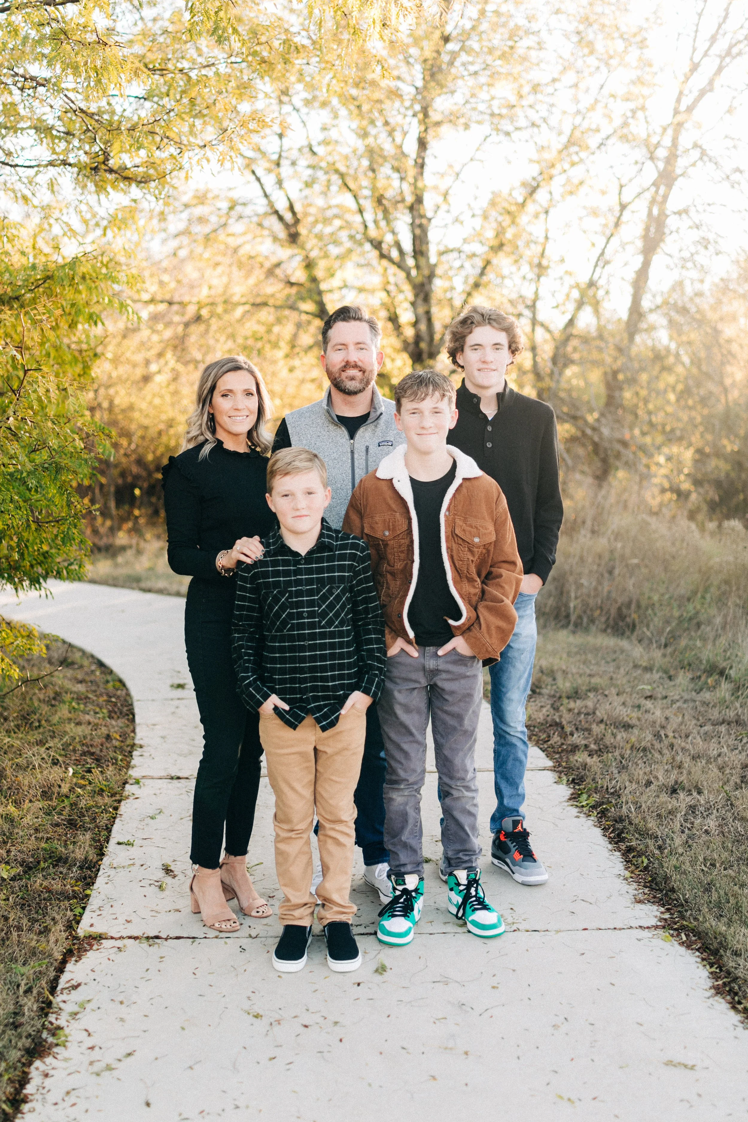 Family of six standing on a sidewalk outdoors during autumn, with trees and golden leaves in the background.
