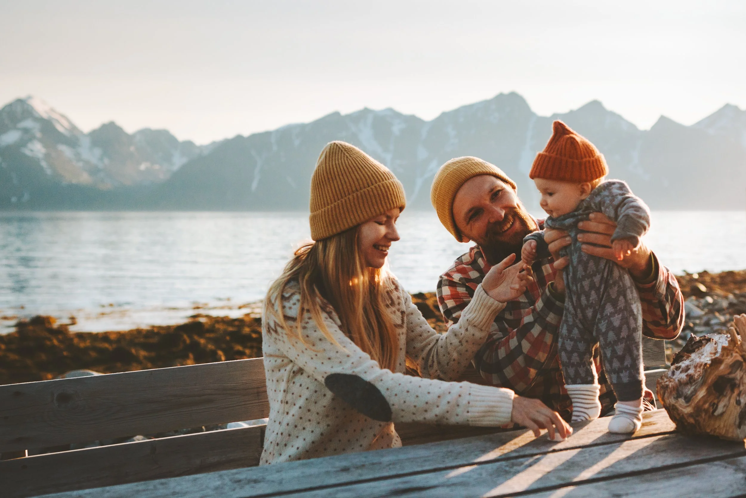 Family of three with a woman, a man, and a baby, enjoying outdoors by a lake with mountains in the background, all dressed warmly with knitted hats.