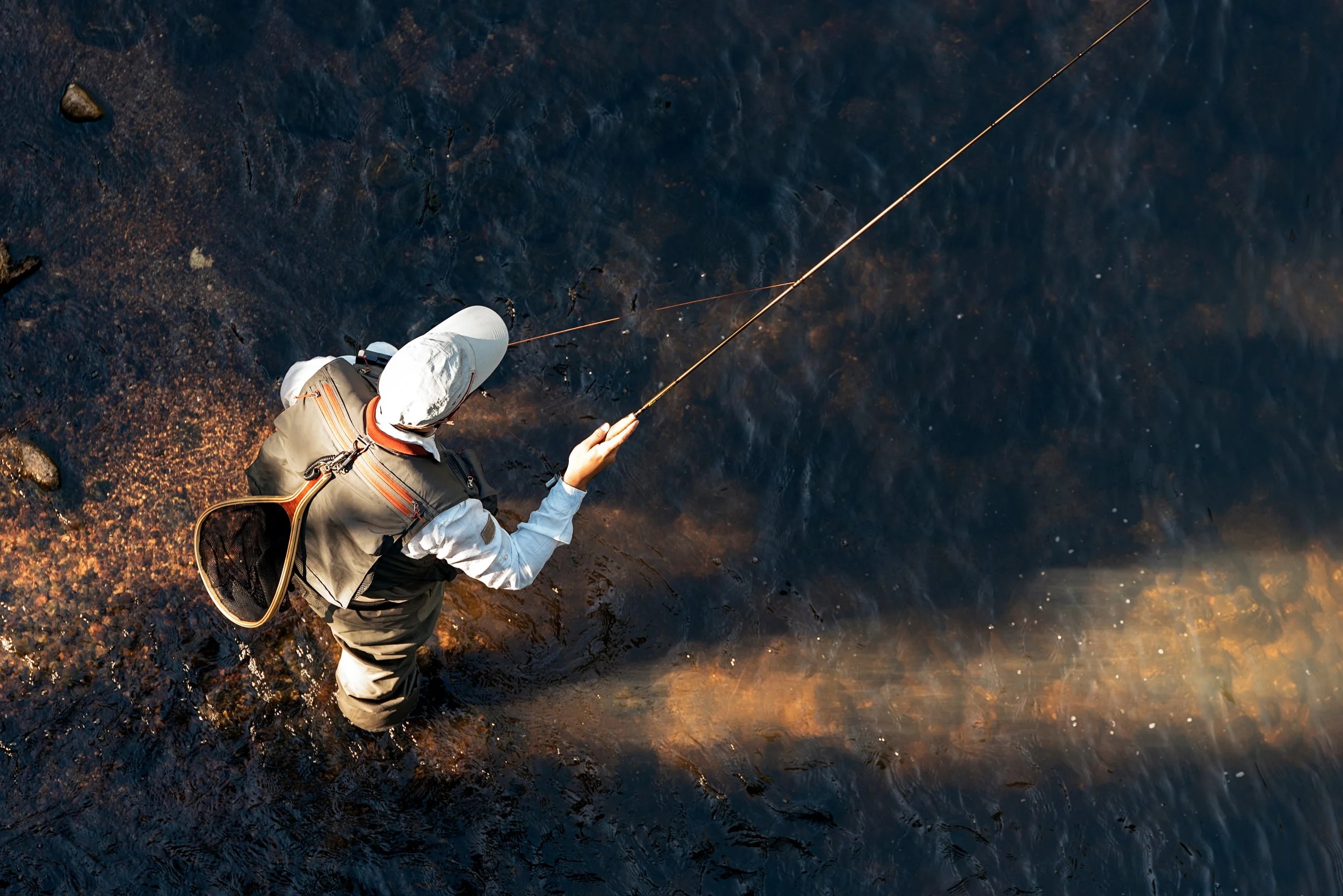 A person standing in shallow water, fishing with a rod, wearing outdoor clothing and a hat.