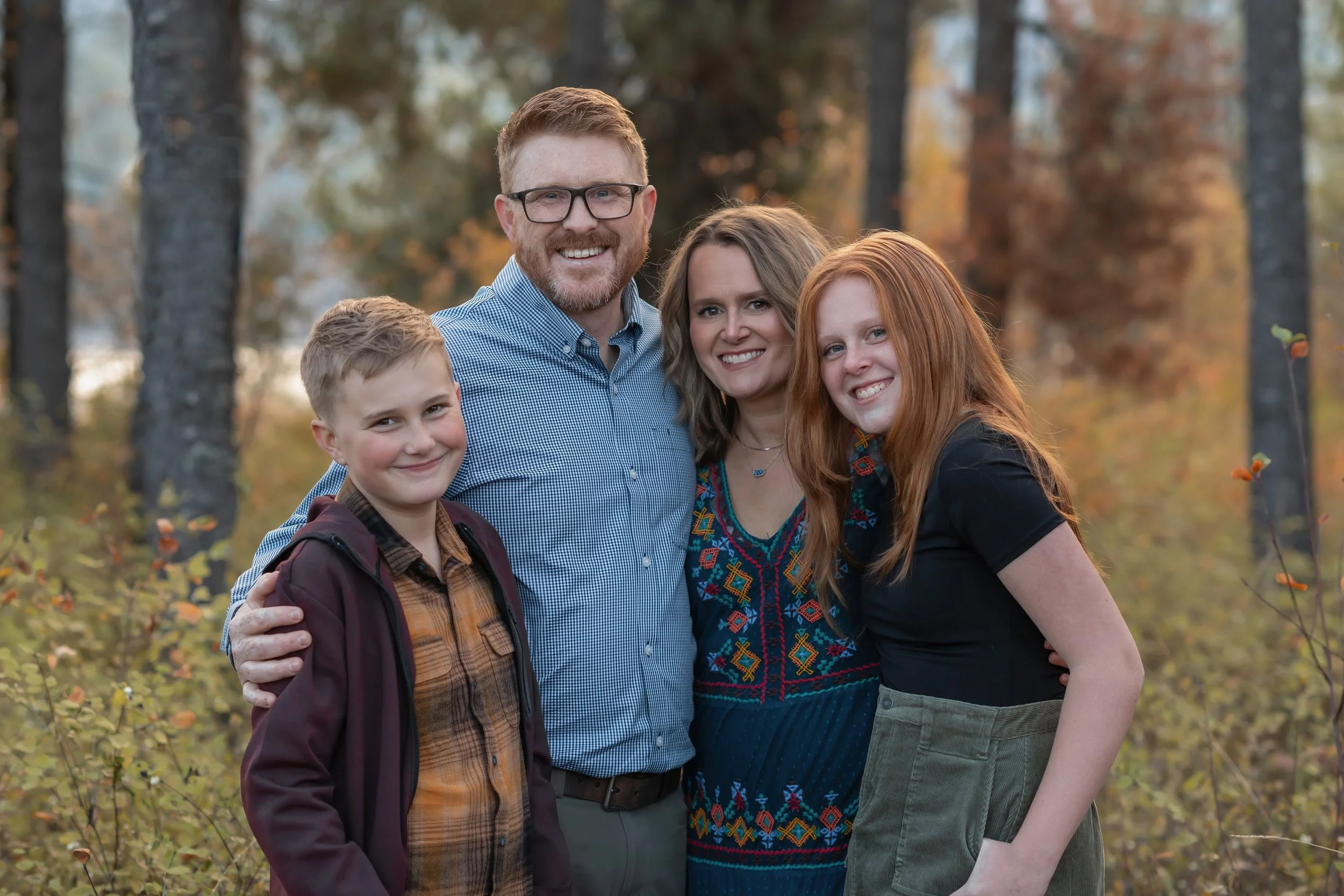 Family of four standing outdoors in a wooded area during fall, smiling at the camera.