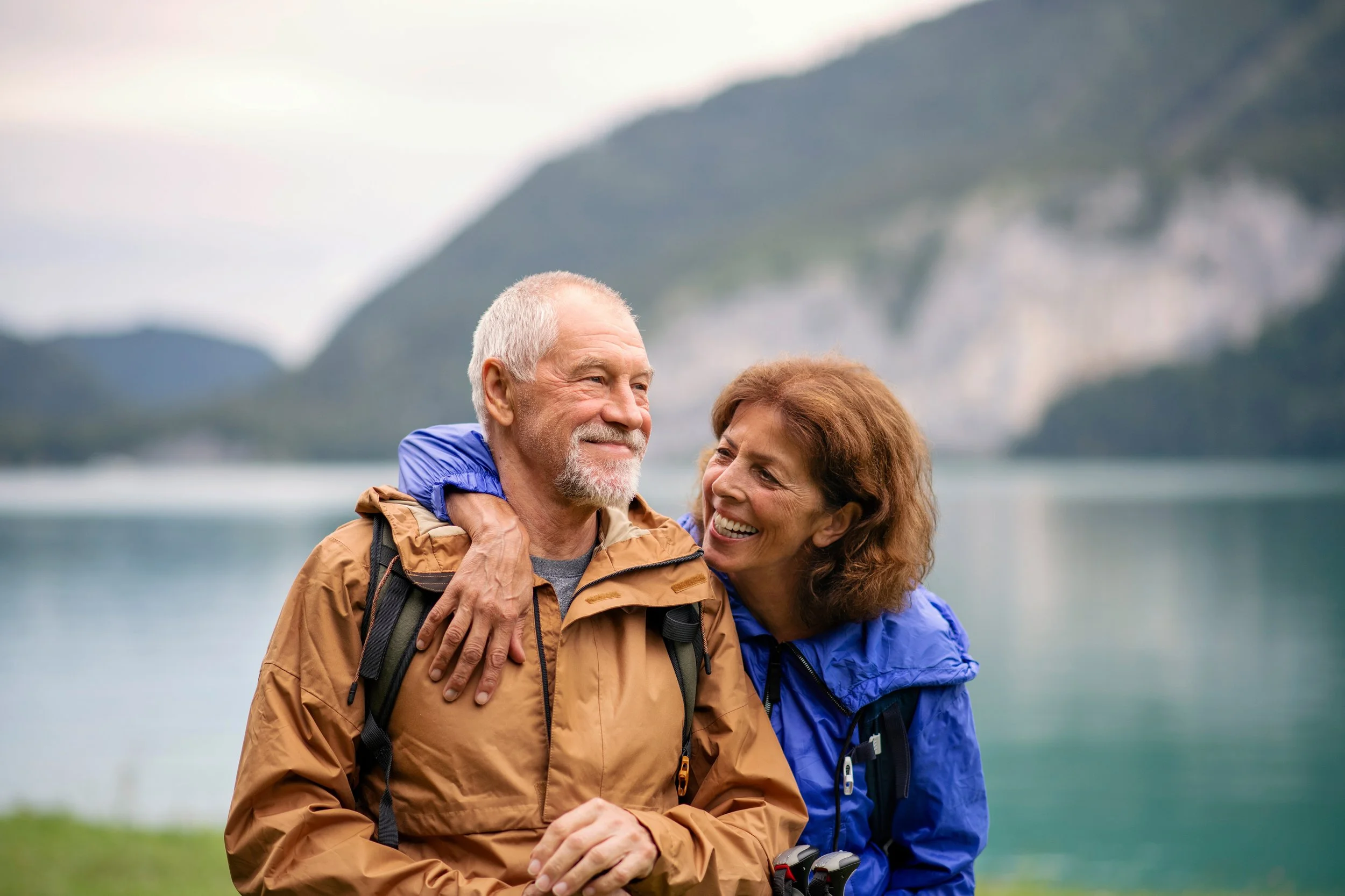 A happy older couple enjoying the outdoors by a lake with mountains in the background. The woman is affectionately hugging the man from the side, both smiling.