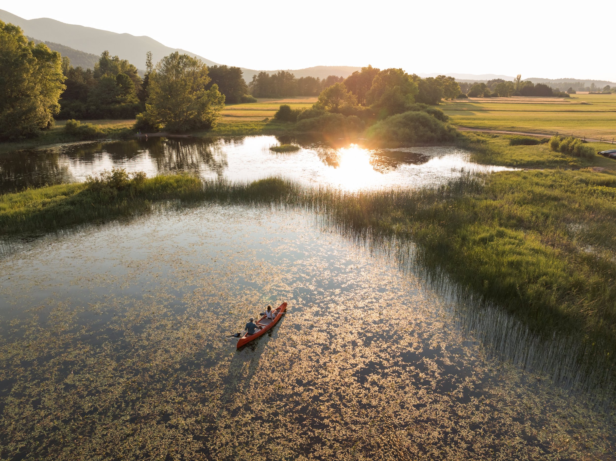 Two people kayaking on a calm pond at sunset with lush green trees, grassy land, and mountains in the background.