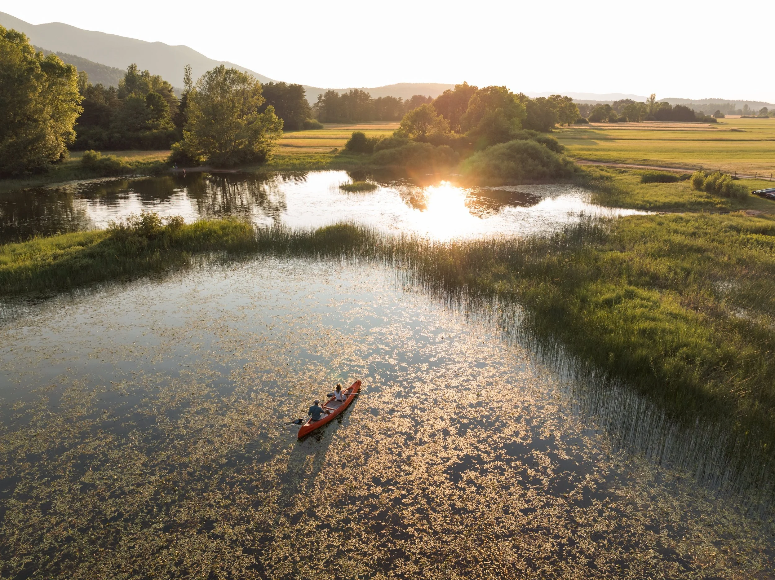 Two people kayaking on a calm pond at sunset with lush green trees, grassy land, and mountains in the background.