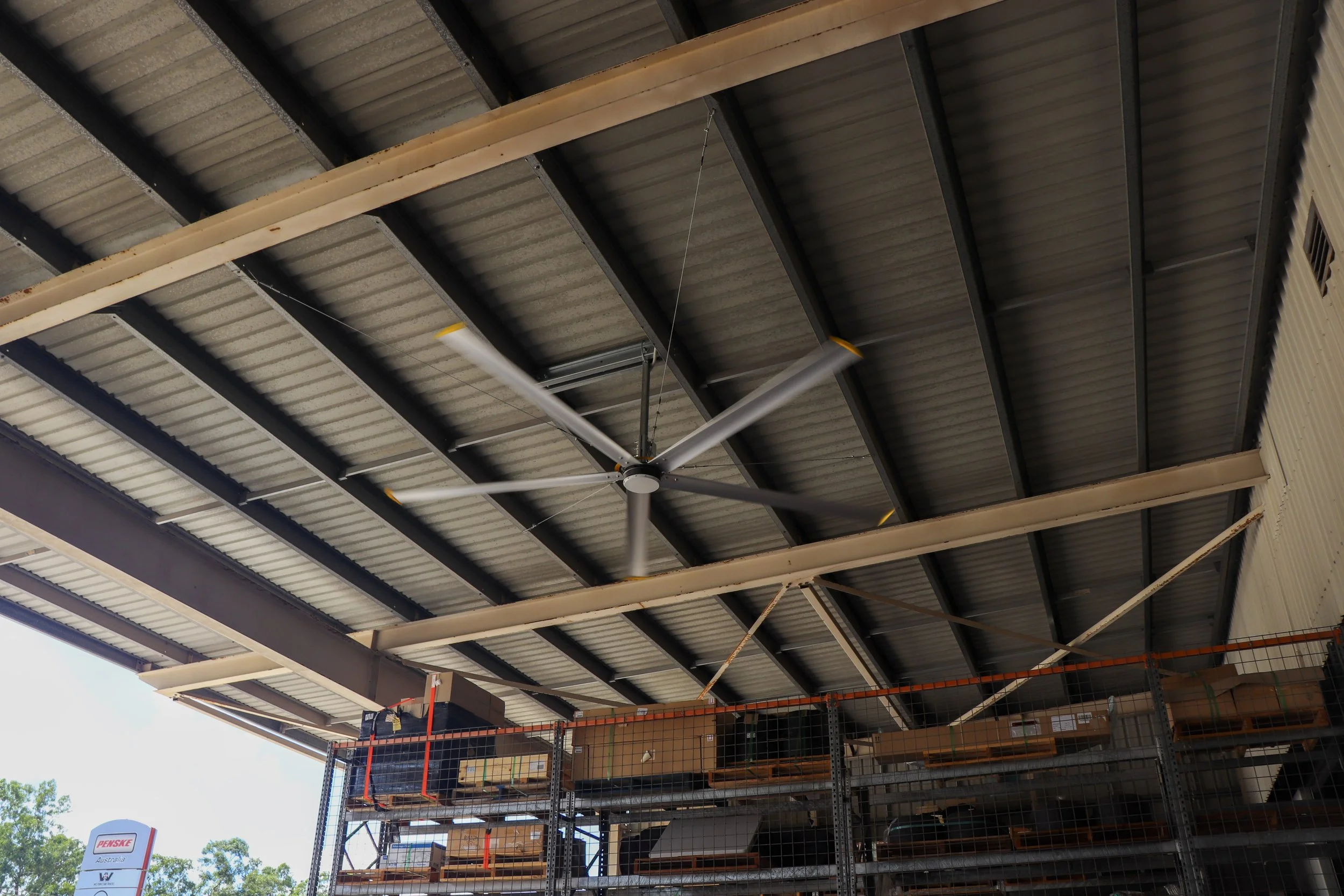 The interior of a warehouse with a corrugated metal roof, a large HVLS big yellow fan, and shelving with boxes and supplies.