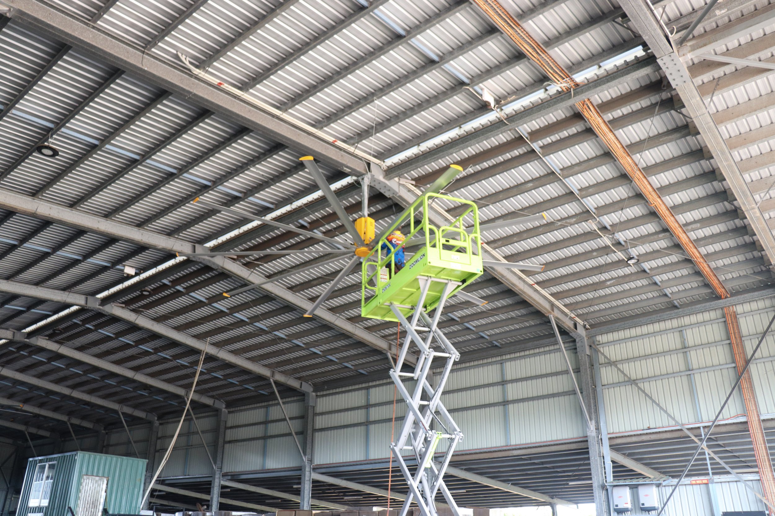 Inside a large industrial warehouse with a high metal ceiling, a green scissor lift is elevated, supporting workers working on the big yellow fan HVLS Top End