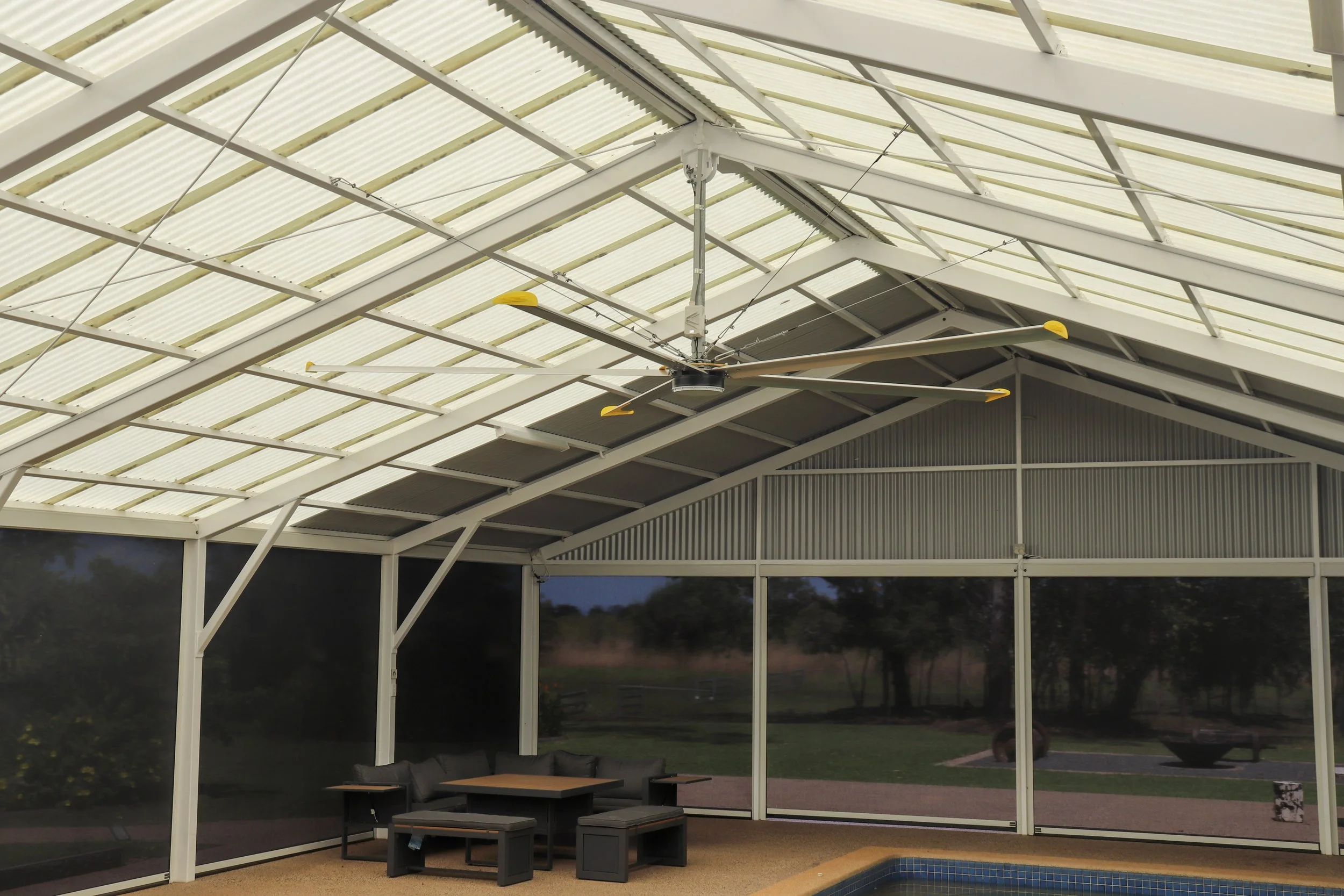 An enclosed Darwin outdoor area with a 3.6m big yellow ceiling fan, patio furniture, and a partial view of a swimming pool outside, under a white metal-framed roof.