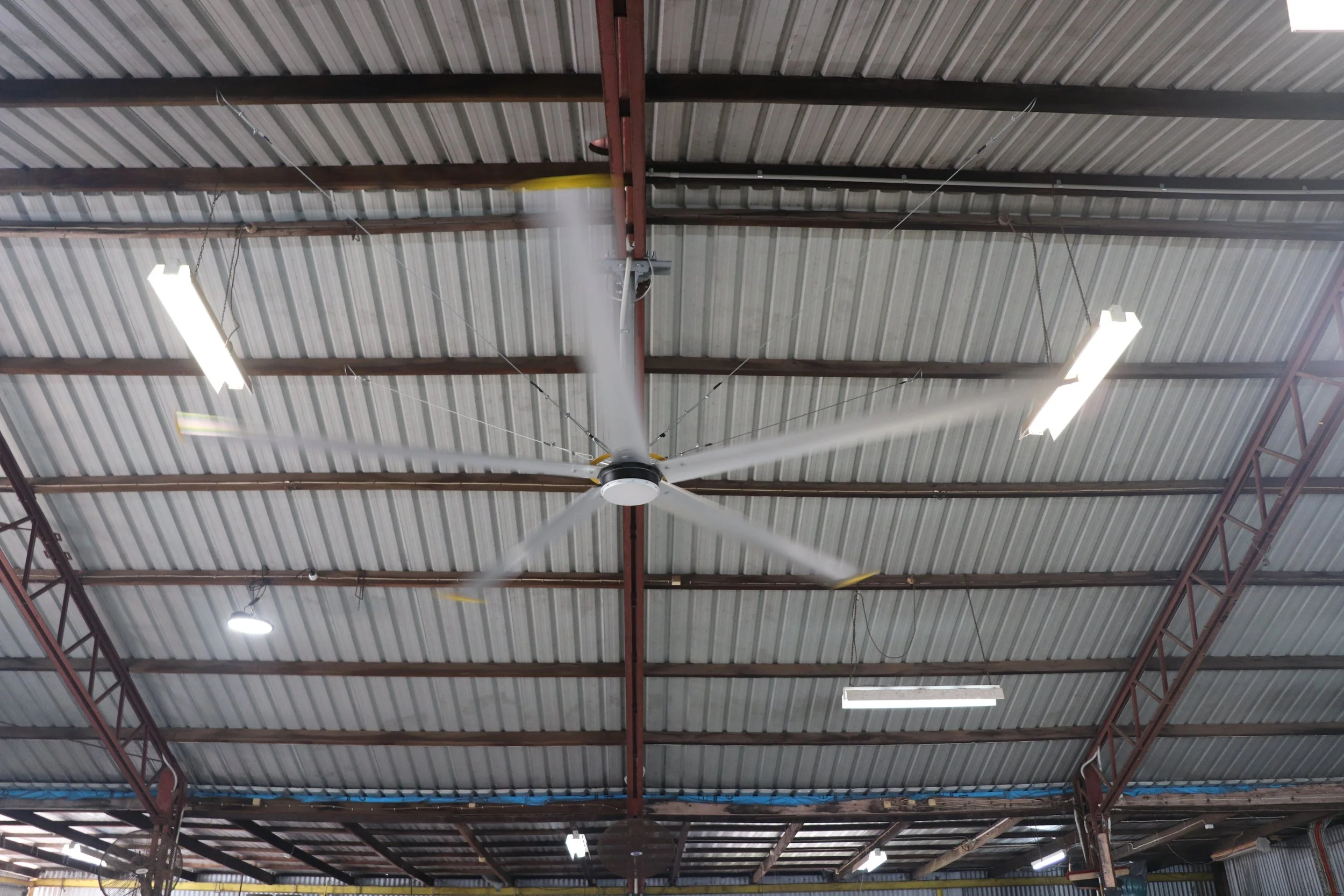View of a warehouse ceiling with metal roofing, red steel beams, fluorescent lights, and a HVLS big yellow ceiling fan.