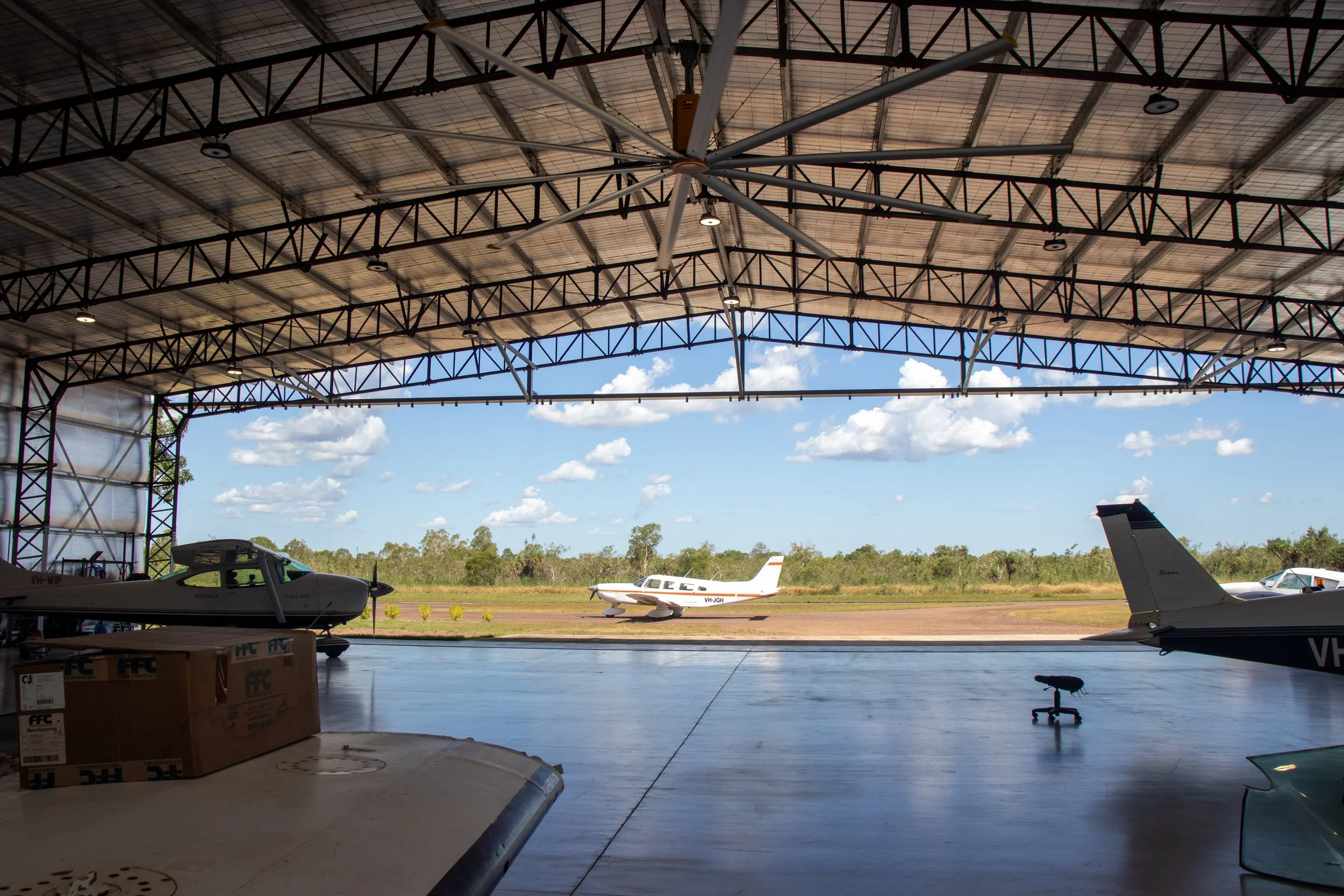 View from inside an airplane hangar showing small private airplanes parked outside on the tarmac with a industrial fan
