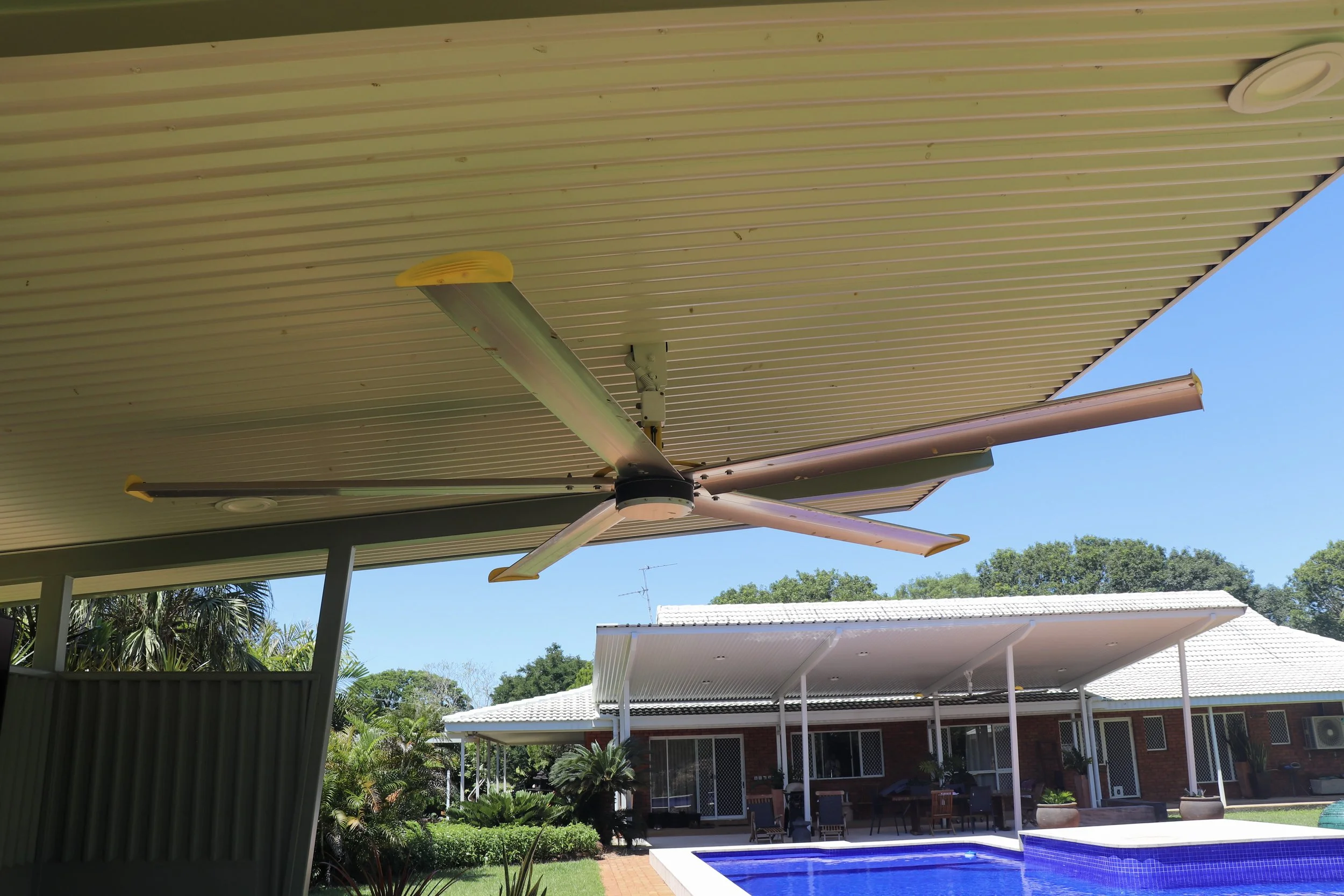 View of a Darwin backyard patio with a swimming pool, outdoor furniture, and covered seating area, under a roof with a small residential big yellow HVLS ceiling fan.