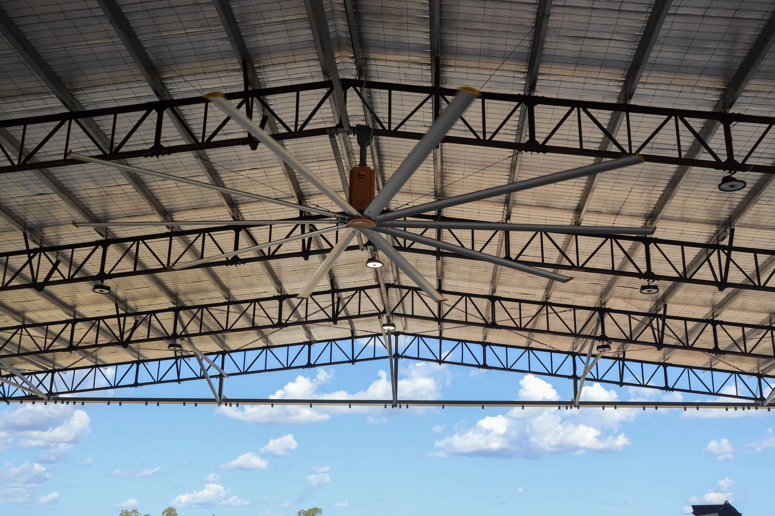 View of a large industrial ceiling with metal trusses, a HVLS big yellow ceiling fan, and solar panels, under a partly cloudy sky.