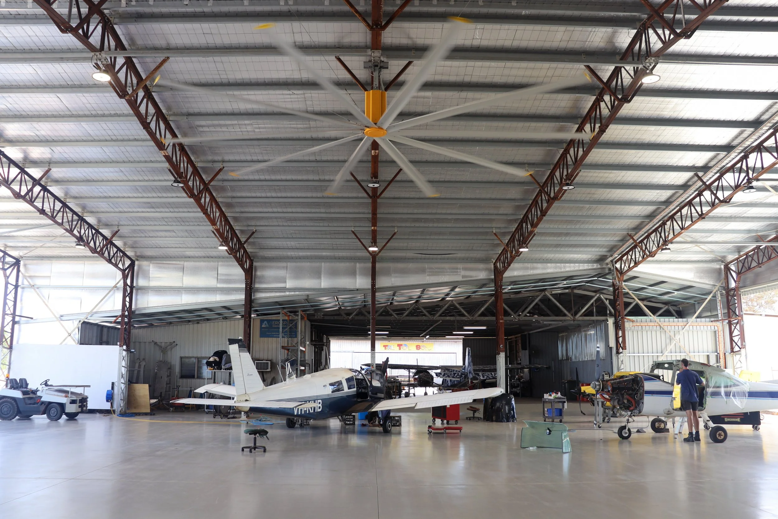 Interior of an aircraft hangar with aircraft, maintenance equipment, and a large 7.2m HVLS big yellow ceiling fan.
