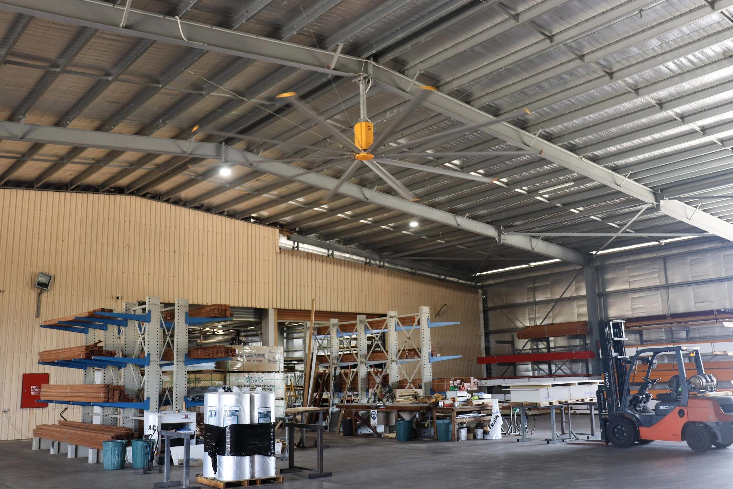 Warehouse or industrial building with metal roof and large big yellow Fan, shelving units with wood and metal materials, construction items, and a forklift.