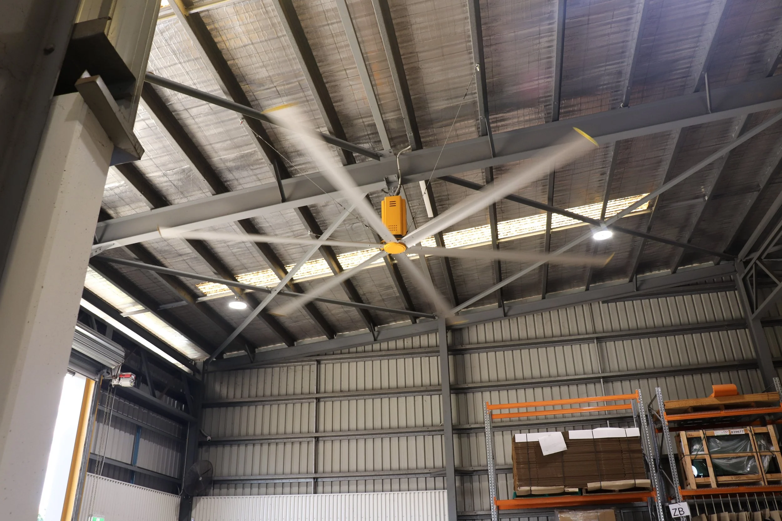 The image shows the interior of a warehouse or industrial building with a HVLS big yellow fan in motion, metal beams, and roofing panels. There are shelves with stacked boxes and items along the lower right side.
