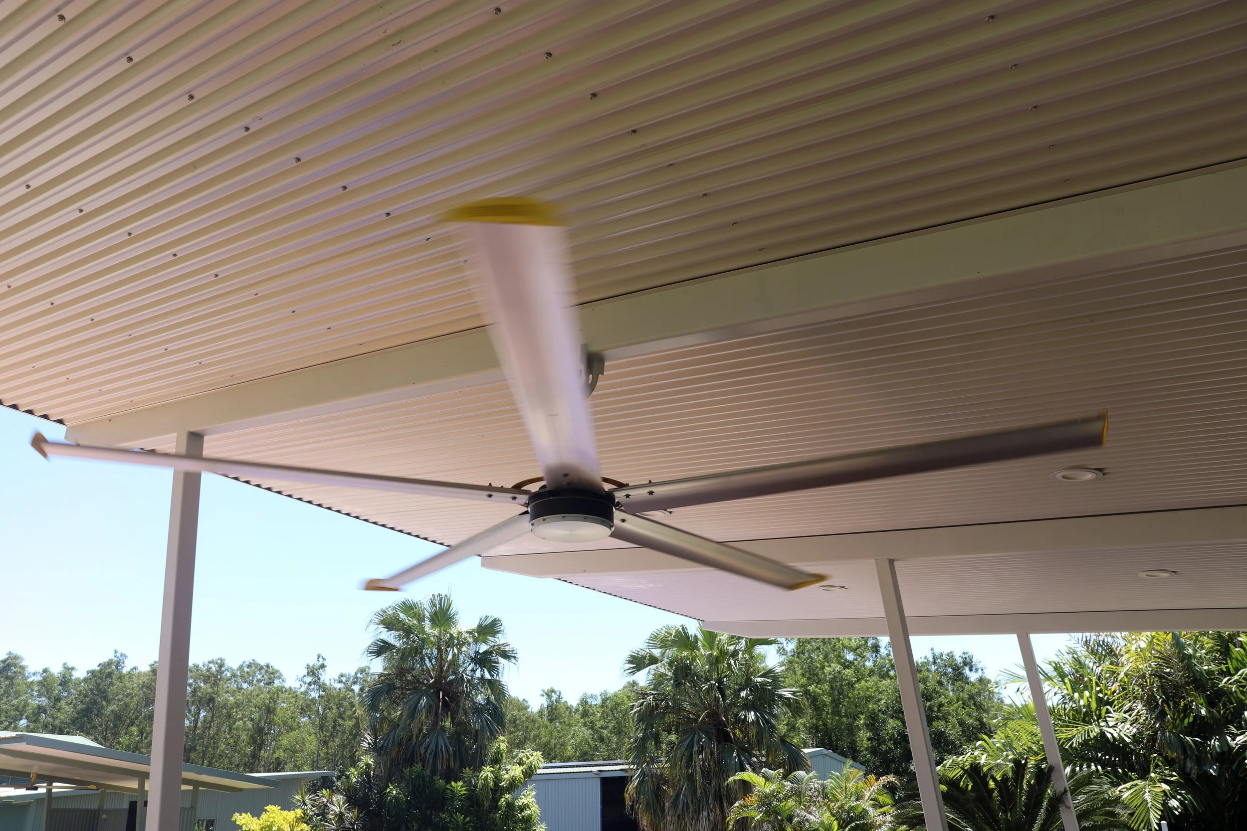 HVLS residential 3.6m ceiling fan with light installed under a beige slatted patio roof, with trees and blue sky in the background.