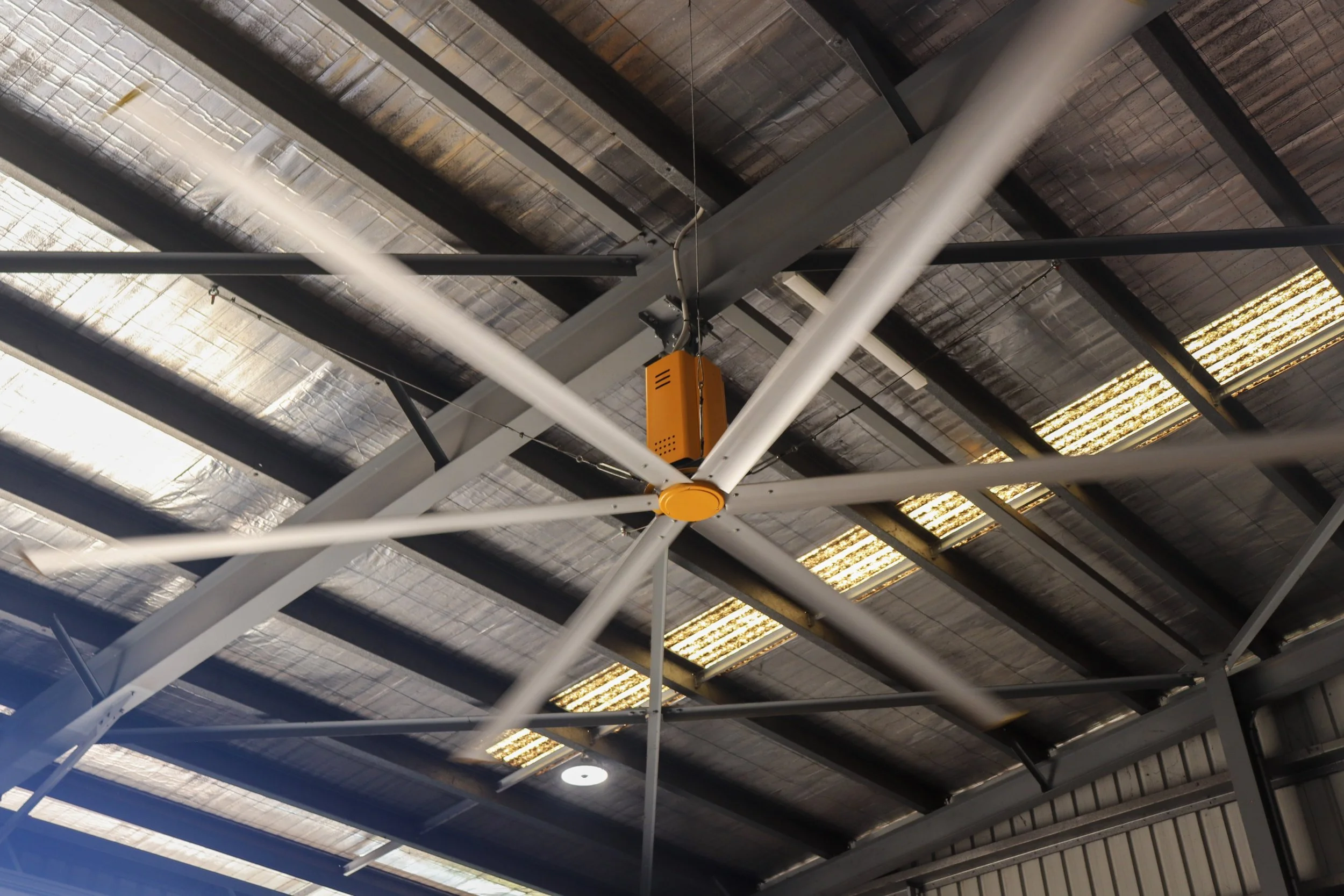 Ceiling of an industrial or commercial building with a large ceiling fan, metal beams, and insulation panels.