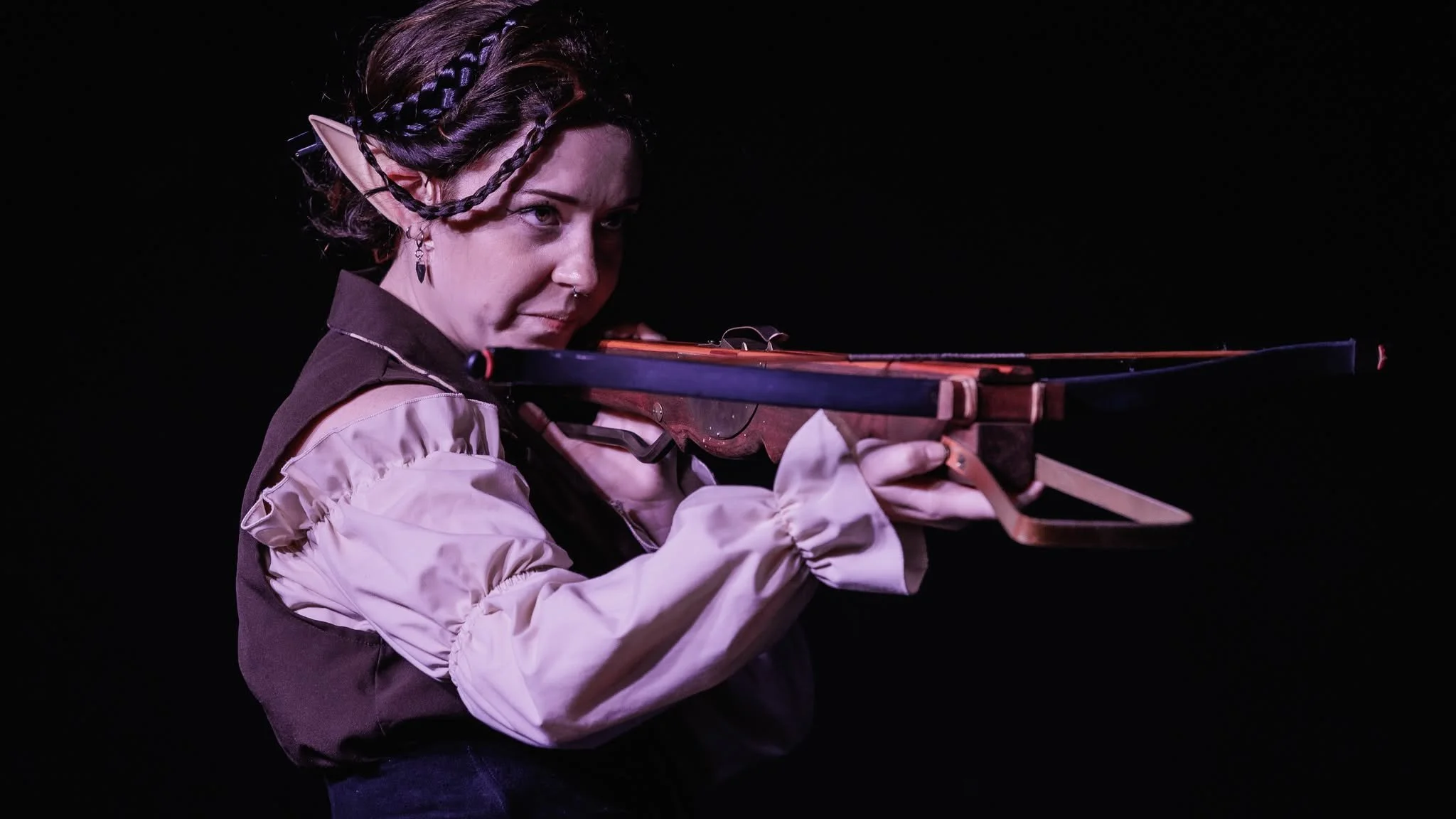 Person in fantasy costume aiming a crossbow, with pointed elf-like ears, styled hair, earrings, and solo dark background.
