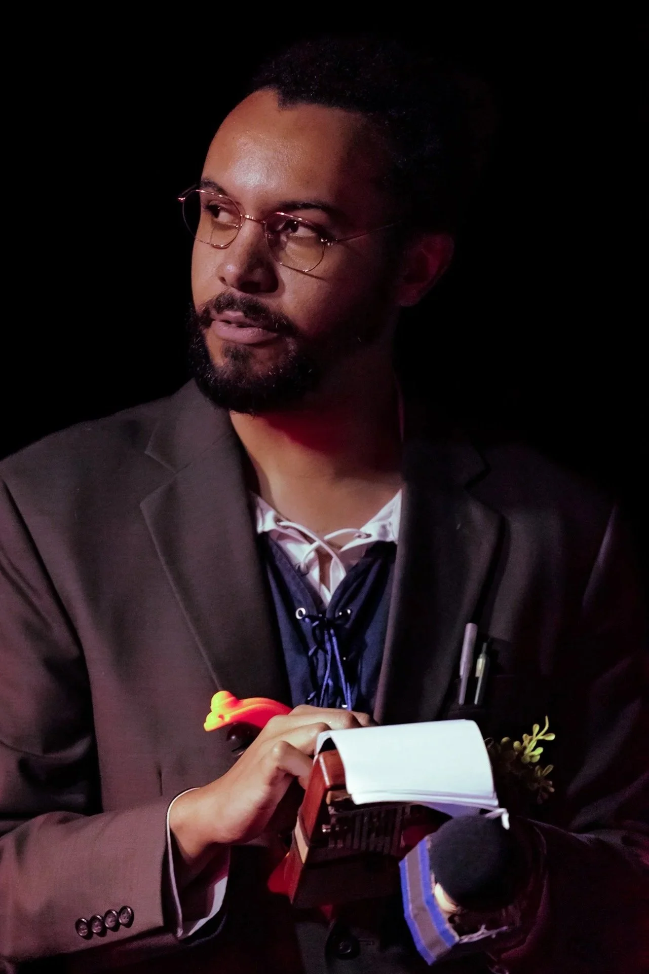 A man in a brown blazer, white shirt, and black tie holding a notepad and pen, with a microphone in front of him, looking to the side against a dark background.