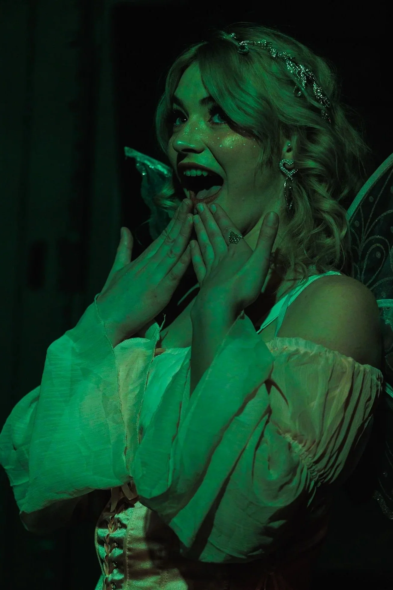 A woman with curly blonde hair, wearing heart-shaped earrings and a metallic headband, expresses surprise or excitement, with hands near her face and mouth open, under green lighting.